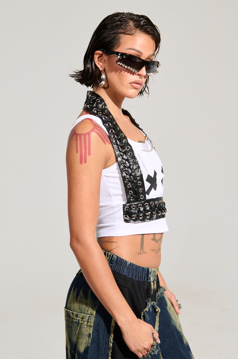 A fashionista with short hair and sunglasses poses confidently in a trendy ensemble. They flaunt the LOCKED DOWN TANK beneath a studded leather vest, paired with distressed jeans. Visible tattoos include a dripping pink heart on the shoulder and other intriguing torso designs.
