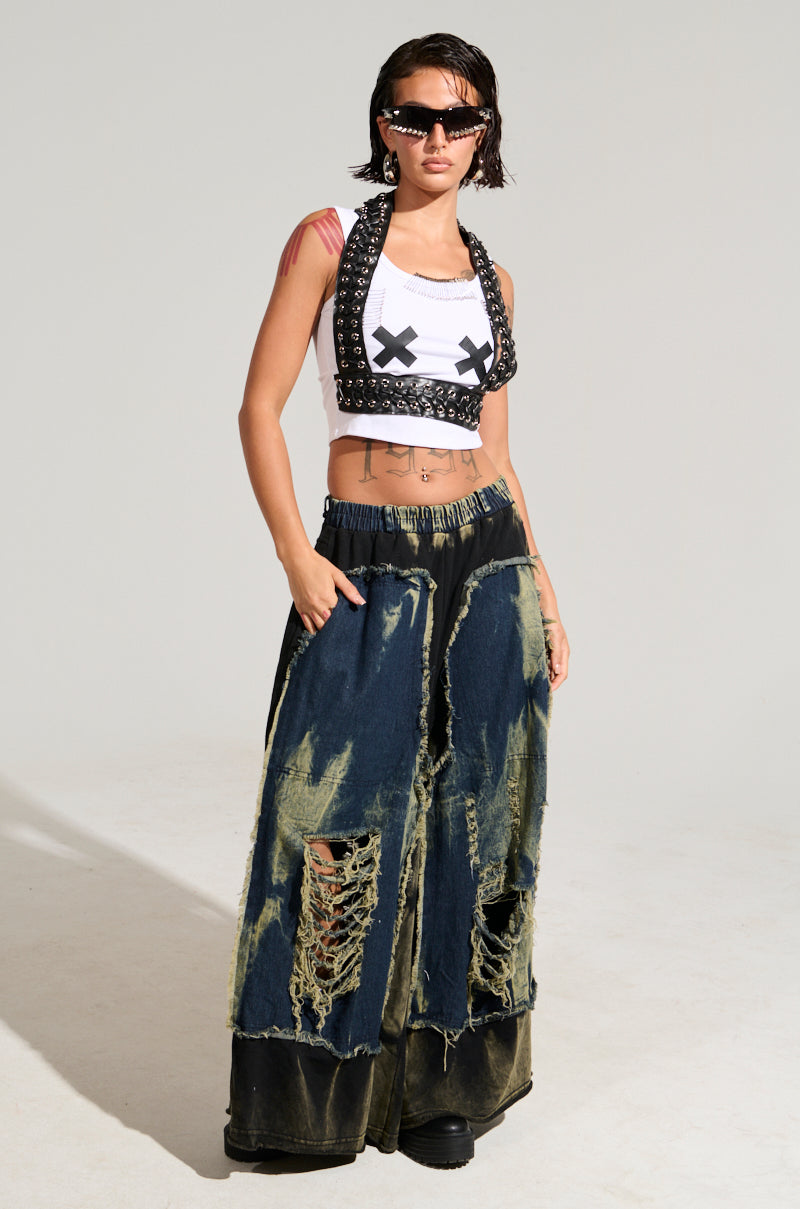 A fashionista exudes confidence in the LOCKED DOWN TANK, paired with large sunglasses and baggy, distressed jeans. The outfit skillfully blends textures and patterns, creating an edgy look against a plain background.