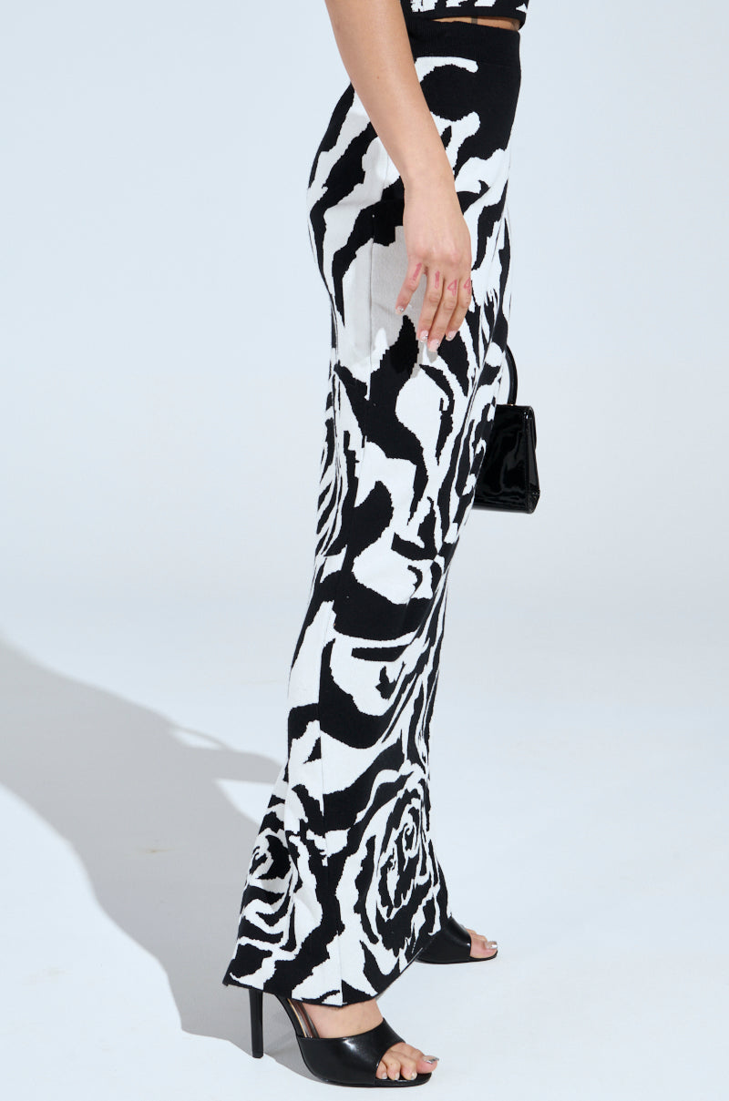 Wearing the ROSEY MAXI KNIT SKIRT, a person stands stylishly in zebra-striped lower wear with black high-heeled sandals, clutching a sleek black accessory. The bright setting highlights their bold look, exuding chic confidence.
