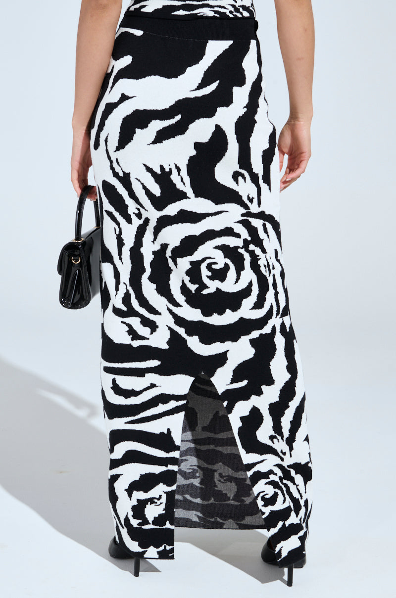 A person is wearing a ROSEY MAXI KNIT SKIRT with a black and white abstract rose print. They pair it with a small black handbag and black heels. The skirt features a high-rise design and a back slit for added flair.