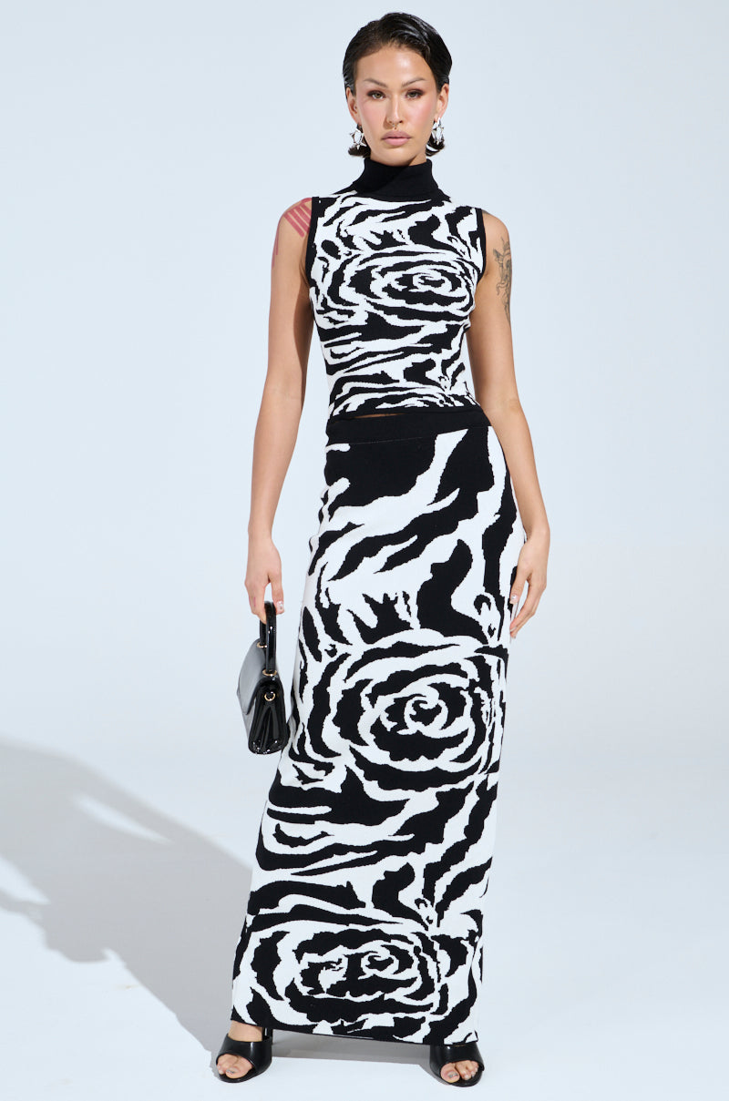 A person stands wearing the sleeveless ROSEY MAXI KNIT SKIRT dress in black and white, featuring a bold floral pattern similar to a B&W rose print. They hold a small black handbag and wear high-heeled sandals. The plain white background enhances the elegance of their ensemble.