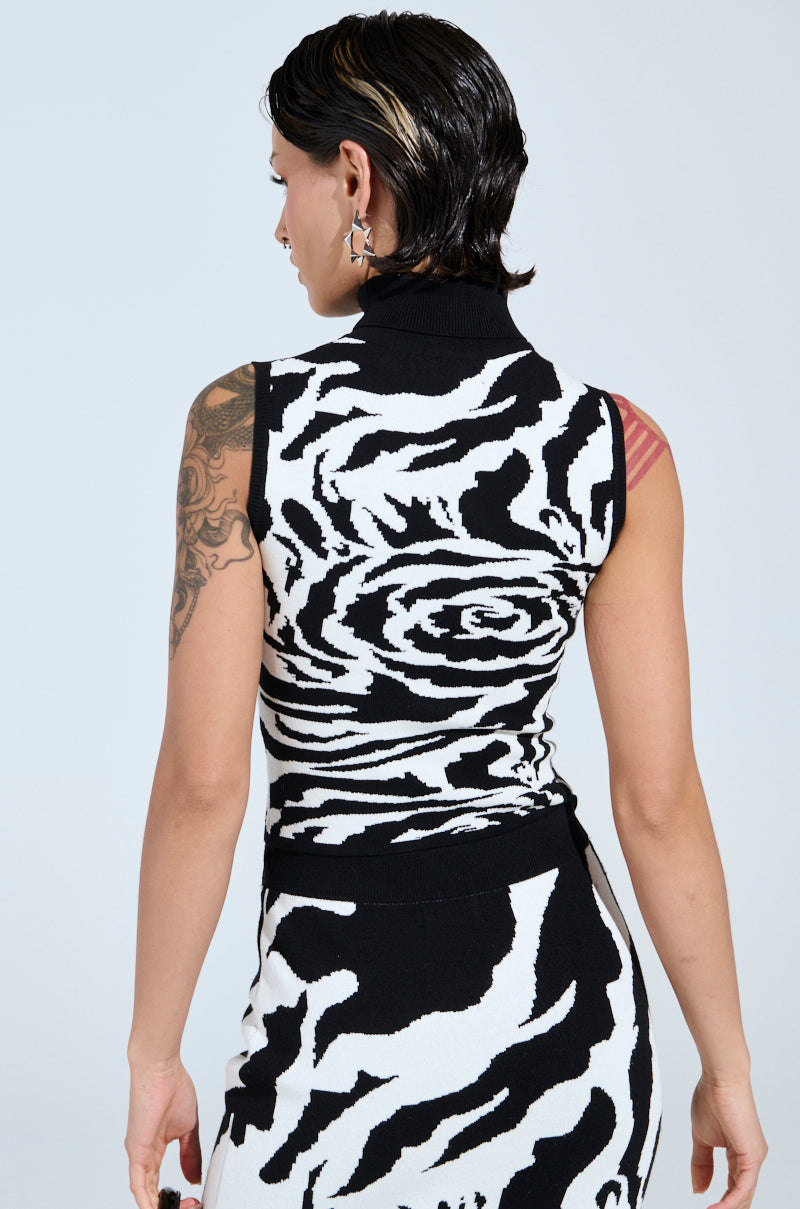 A person with short dark hair faces away, wearing a sleeveless ROSEY MOCK NECK KNIT TOP with a B&W rose print and tattoos on their arms. They accessorize with star-shaped earrings against a plain, light background, capturing the chic essence of an AKIRA Label ensemble.