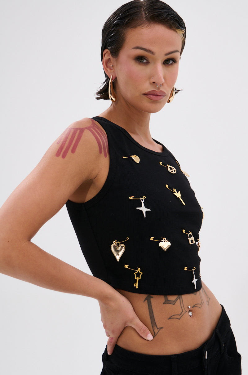 A woman with slicked-back dark hair wears the LOVIN' YOU EMBELLISHED TANK TOP featuring gold charms and safety pins. She accessorizes with hoop earrings, a nose ring, and tattoos, standing against a plain background. 0