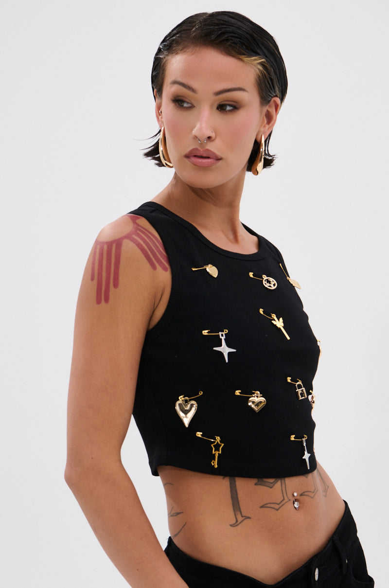 Someone with short, slicked-back hair wears the LOVIN' YOU EMBELLISHED TANK TOP, featuring gold safety pins and charms. They also sport large hoop earrings, tattoos, and a bold red paint-like mark on their upper left arm. 1