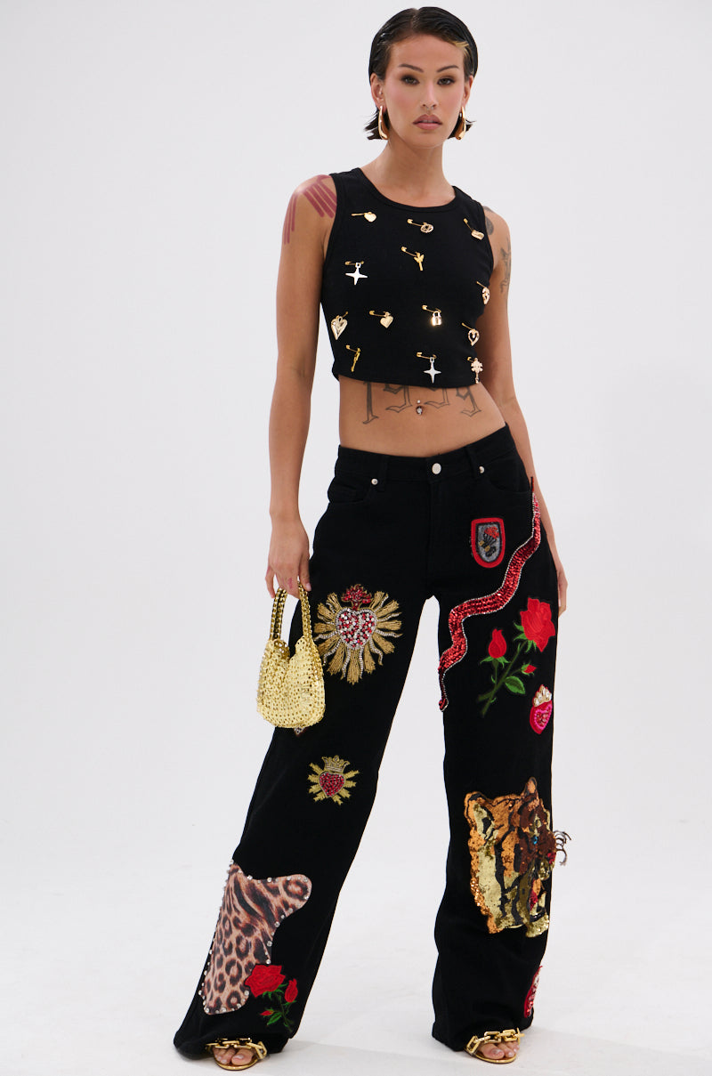 A woman stands confidently in the LOVIN' YOU EMBELLISHED TANK TOP with gold details, paired with black pants featuring colorful embroidery. She finishes the chic look with a small gold beaded handbag. 4