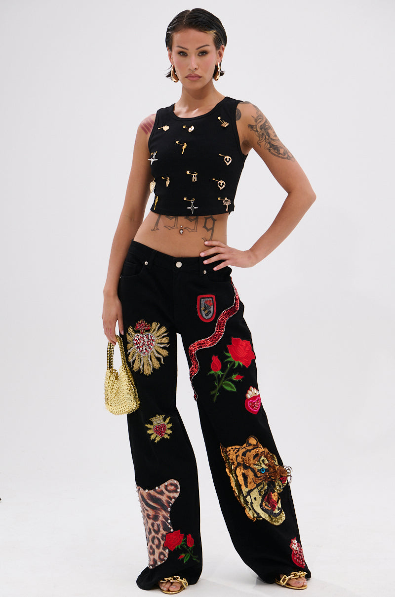 A woman poses confidently in the LOVIN' YOU EMBELLISHED TANK TOP with small gold charms, paired with black wide-leg pants featuring colorful embroidered animal and floral designs. She accessorizes with a small gold purse and displays her tattoos. 5
