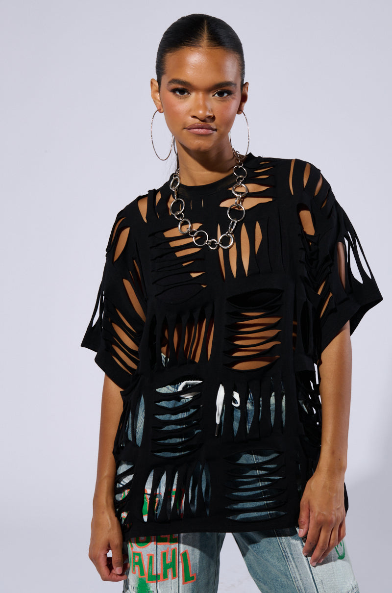 A person wearing the CUT HIM OFF SHORT SLEEVE DISTRESSED T-SHIRT IN BLACK, featuring an oversized fit and a cut-out grid pattern, paired with jeans. They have a sleek hairstyle, large hoop earrings, and a chunky silver necklace. The background is a plain, light grey.