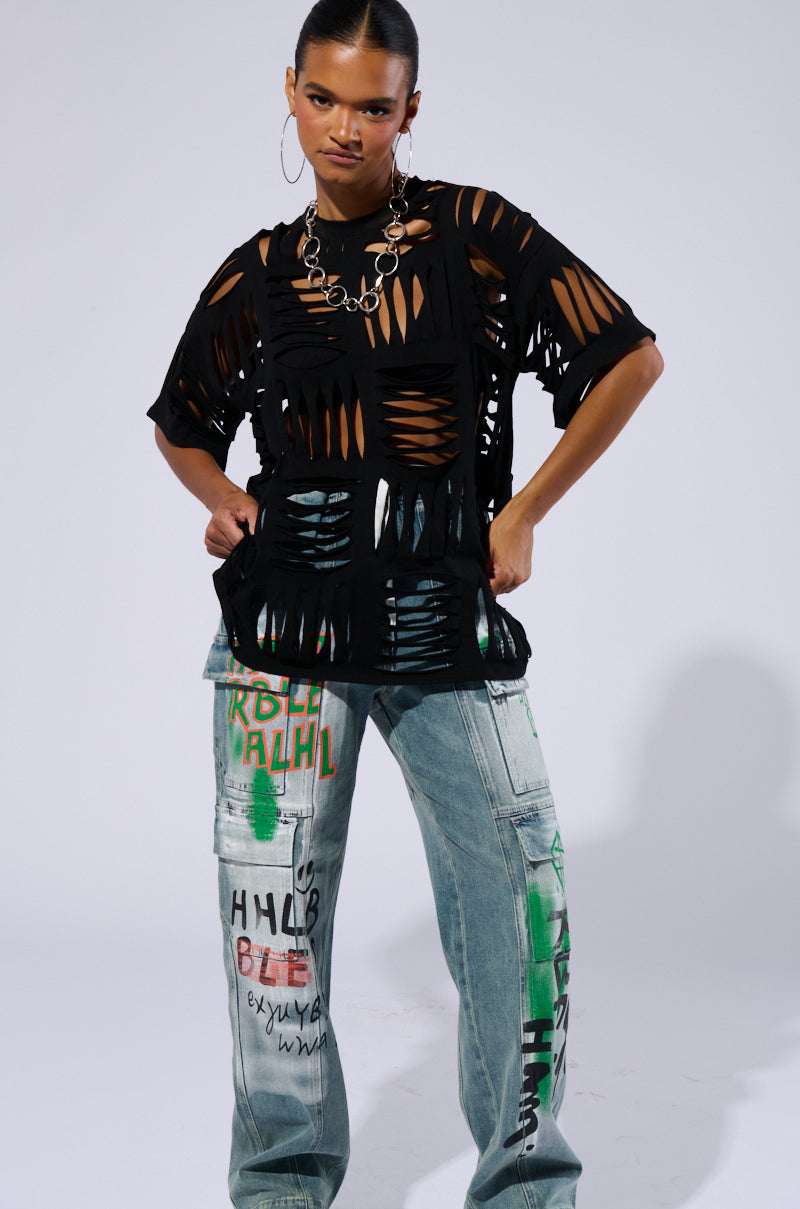 A person stands confidently in the CUT HIM OFF SHORT SLEEVE DISTRESSED T-SHIRT IN BLACK, paired with loose blue jeans decorated with vibrant graffiti. With slicked-back hair, large hoop earrings, and a chunky chain necklace, they pose with hands on hips against a neutral background.