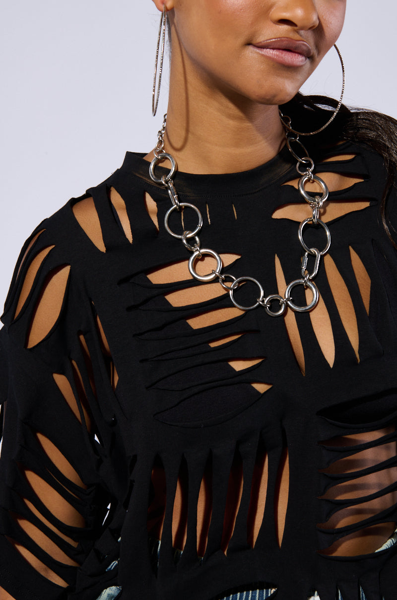 A person wearing the "CUT HIM OFF SHORT SLEEVE DISTRESSED T-SHIRT IN BLACK" featuring a cut-out grid pattern and a chunky silver chain necklace. They also have large, circular hoop earrings. The background is plain and neutral, emphasizing the outfit.