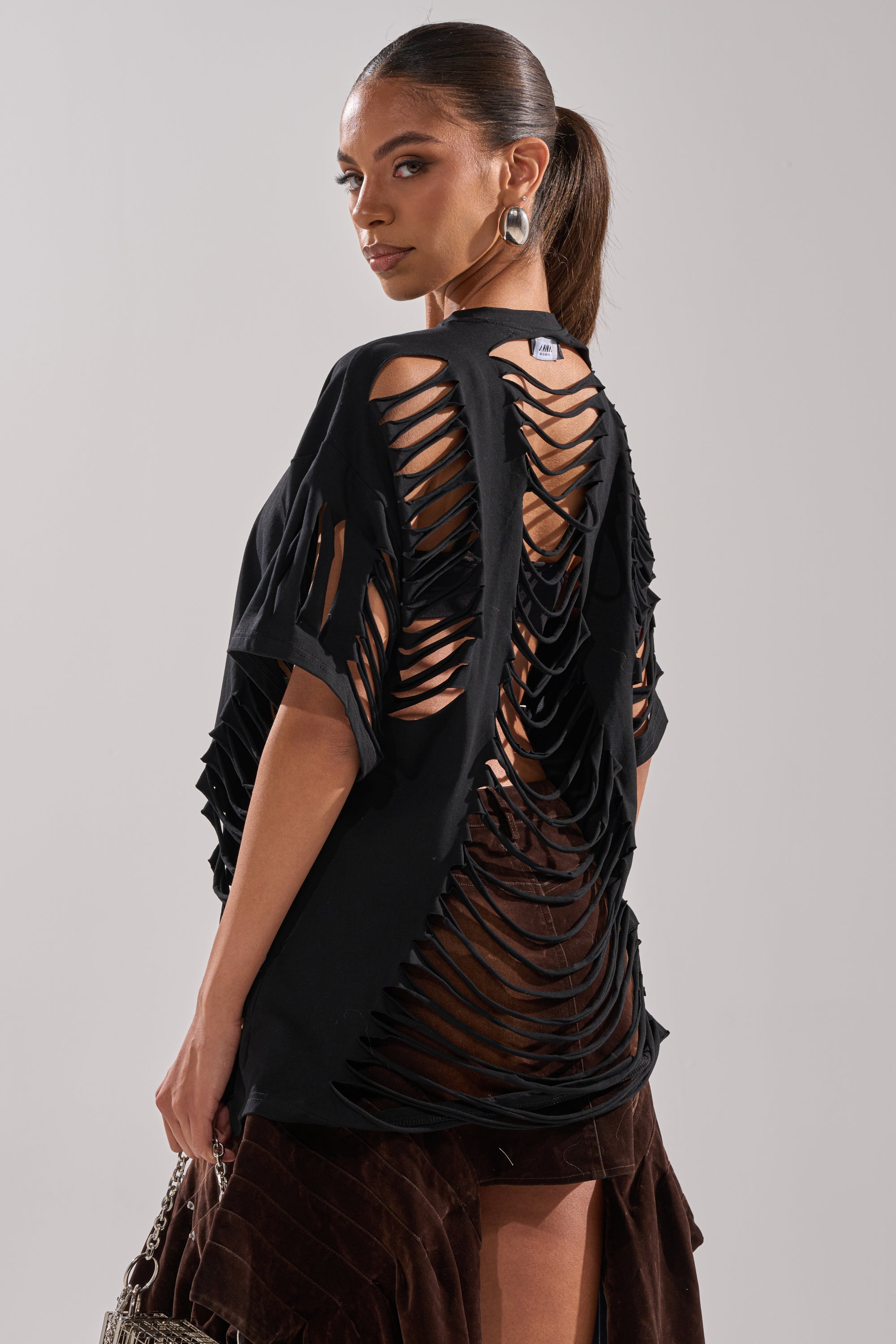 A woman with a sleek ponytail wears the SLASHED UP OVERSIZED SHORT SLEEVE TEE IN BLACK featuring bold slashed cutouts on the back and sleeves, styled with a brown skirt. She glances over her shoulder and holds a textured handbag. 0
