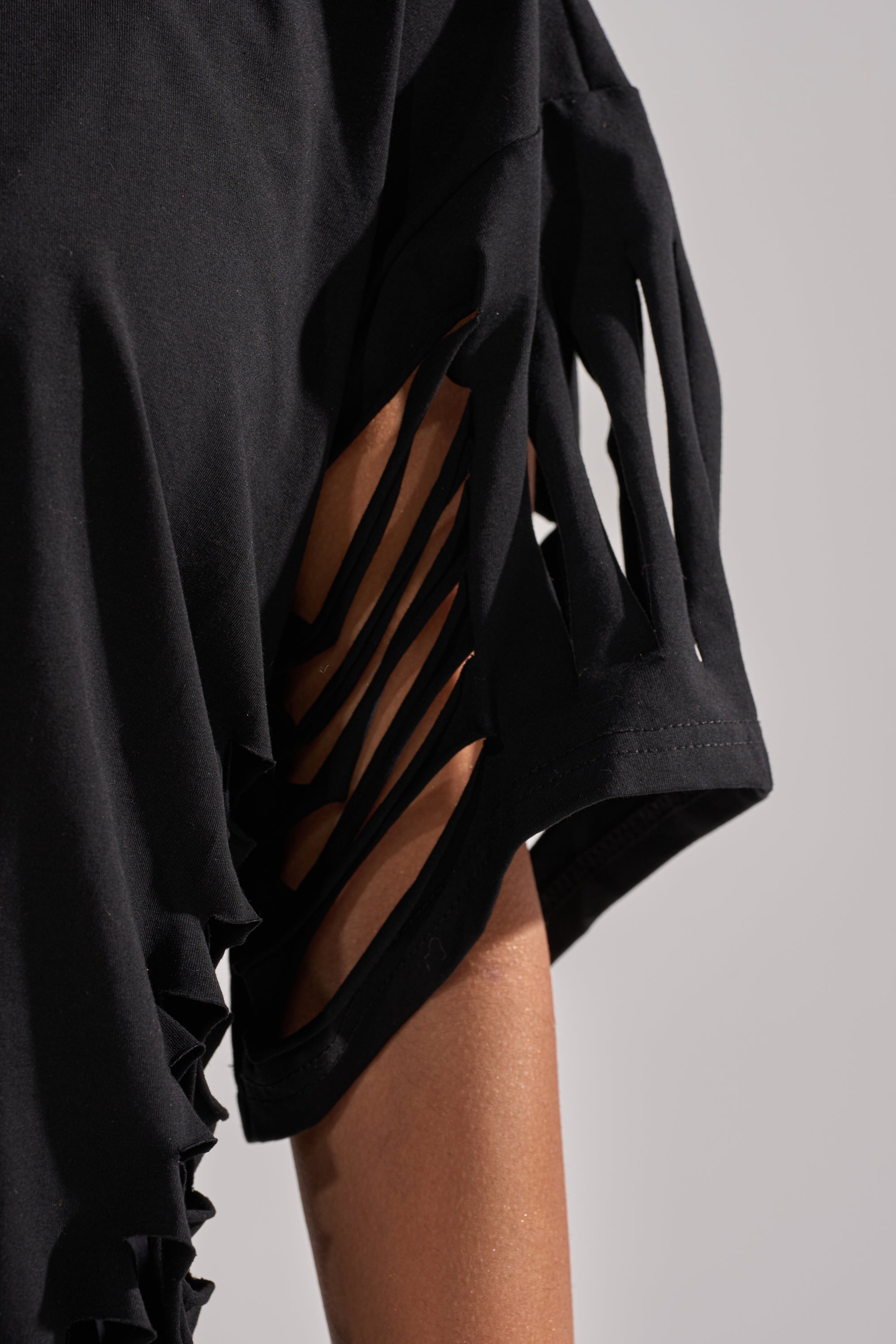Close-up of the SLASHED UP OVERSIZED SHORT SLEEVE TEE IN BLACK, featuring multiple sleeve slits that reveal the wearer’s arm. This bold statement top stands out against a plain, neutral background. 9