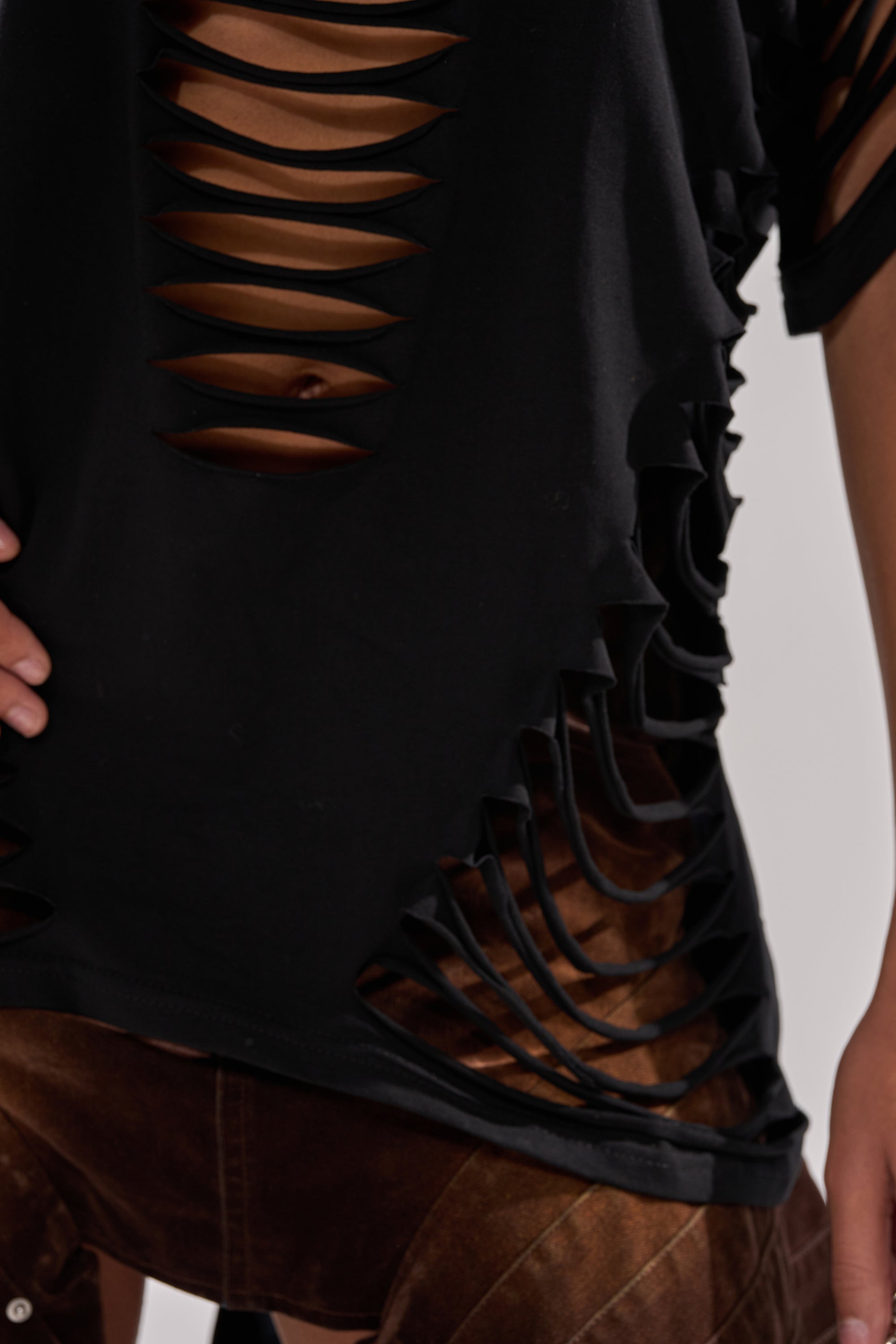 Wearing the SLASHED UP OVERSIZED SHORT SLEEVE TEE IN BLACK with multiple horizontal slits, this person pairs it with brown pants. Only the torso and part of one arm resting on their hip are visible. 10