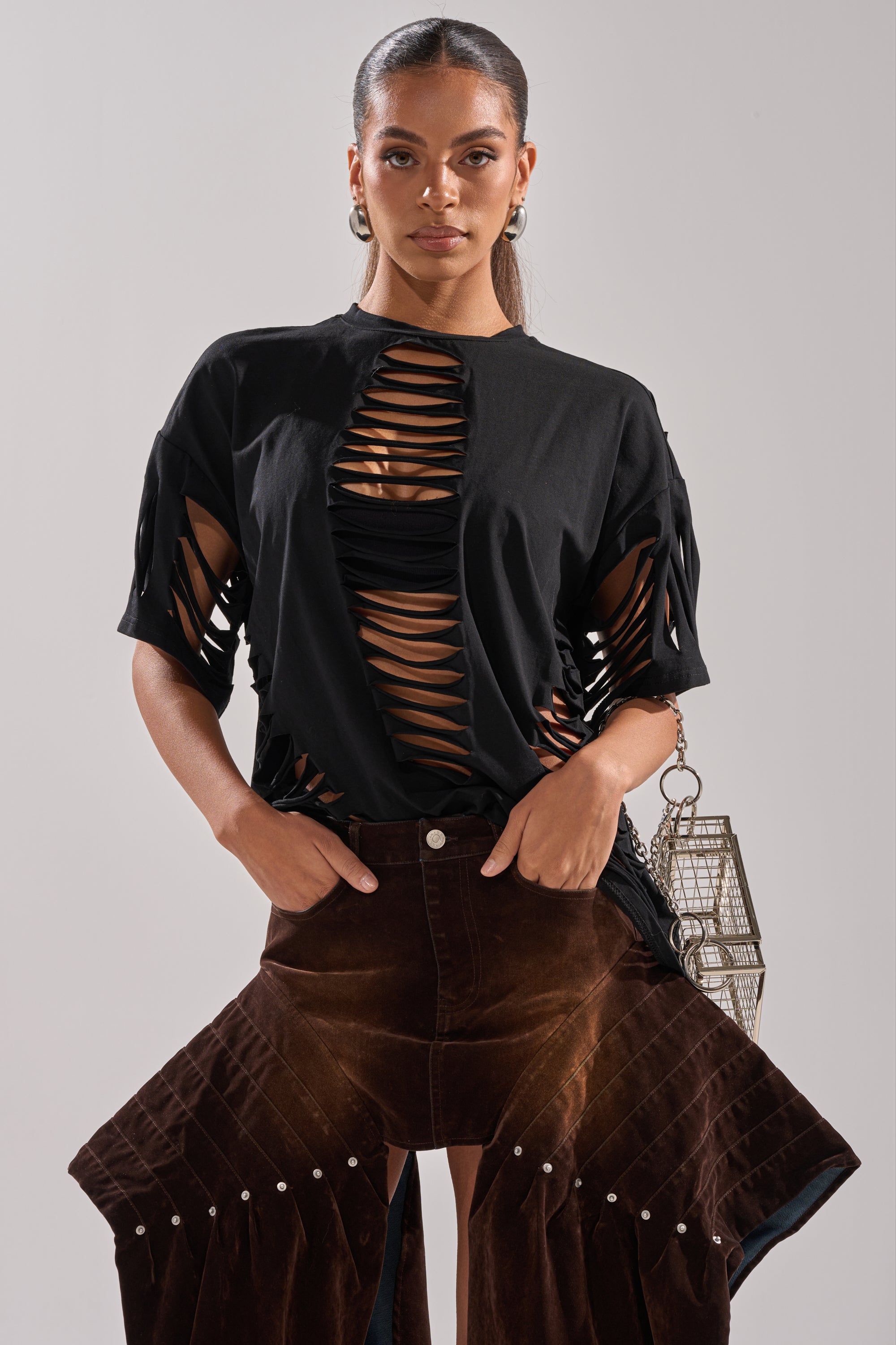 A woman stands confidently in an AKIRA Label SLASHED UP OVERSIZED SHORT SLEEVE TEE IN BLACK and high-waisted brown wide-leg pants. Her hair is pulled back, paired with hoop earrings and a metallic handbag. 1