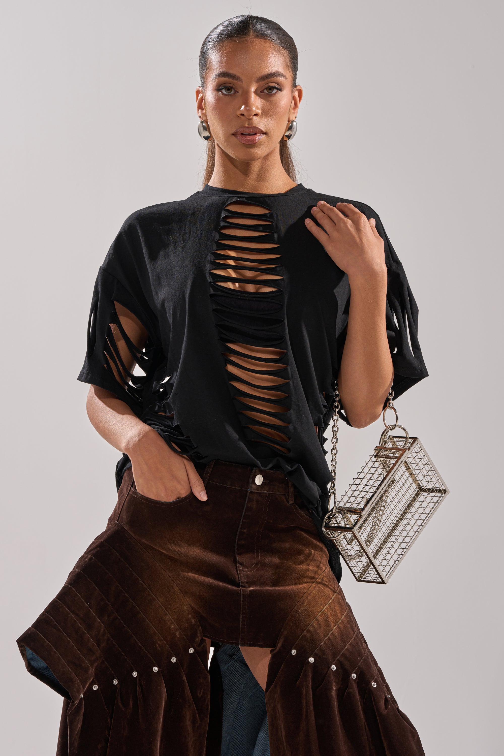 A woman models the SLASHED UP OVERSIZED SHORT SLEEVE TEE IN BLACK, styled with a brown suede pleated skirt and geometric clear handbag. Hoop earrings, sleek hair, and a confident look complete her bold outfit. 3