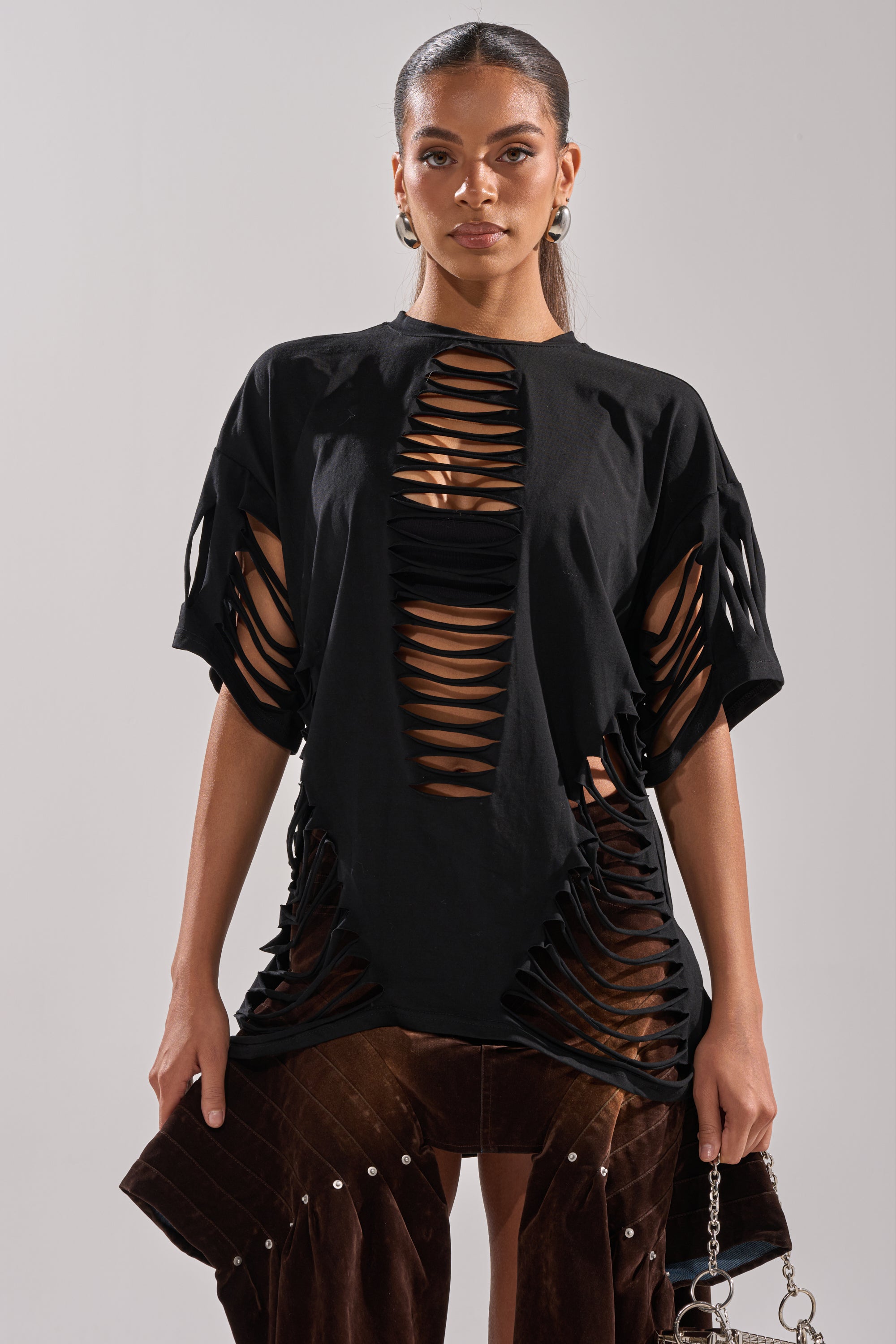 A woman faces forward in the SLASHED UP OVERSIZED SHORT SLEEVE TEE IN BLACK with horizontal slashes, brown pants, a chain-strap bag, pulled-back hair, and hoop earrings for a bold look. 4