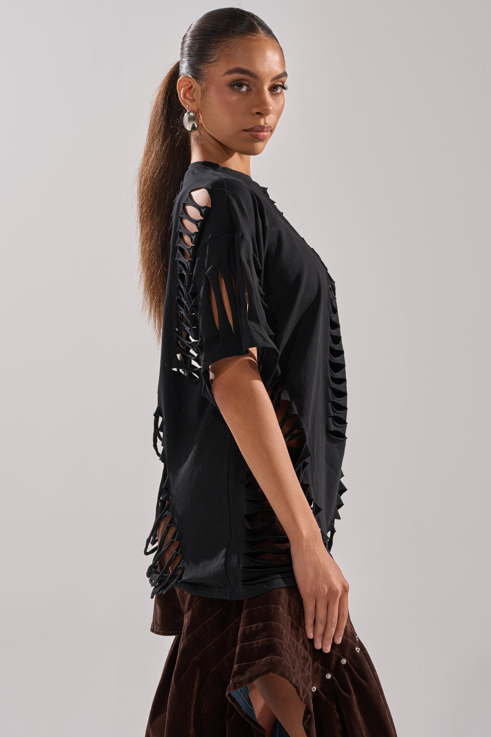 A woman with long, straight hair in a ponytail models the SLASHED UP OVERSIZED SHORT SLEEVE TEE IN BLACK—an edgy top with multiple slashes—paired with brown pants, posing sideways against a plain light gray background. 5