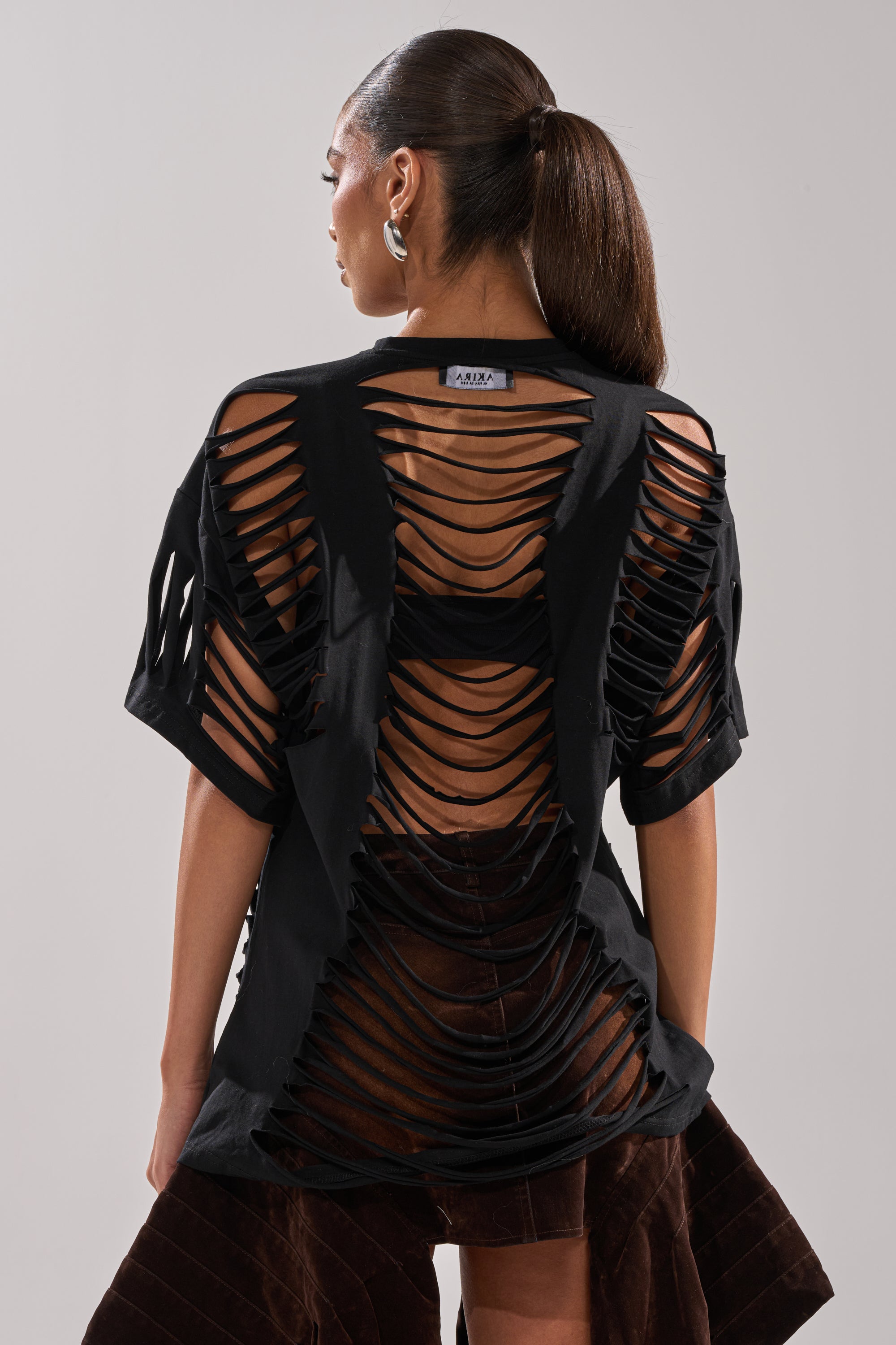 A woman with a sleek ponytail wears the SLASHED UP OVERSIZED SHORT SLEEVE TEE IN BLACK, its cut-outs revealing a black undergarment. She accessorizes with large hoop earrings and a dark brown skirt. 6