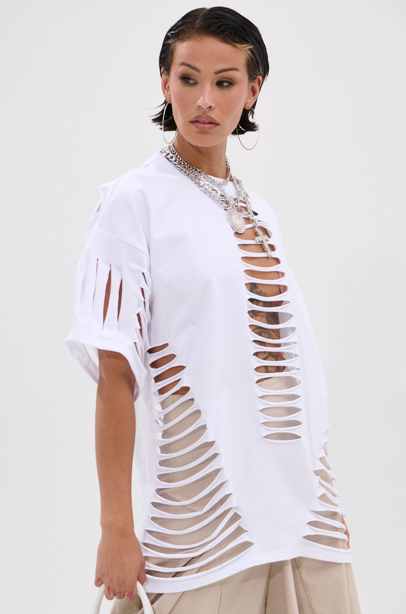Wearing the SLASHED UP OVERSIZED SHORT SLEEVE TEE with bold horizontal cutouts over beige pants, this look is finished with hoop earrings, layered necklaces, and slicked-back hair against a simple background. 0