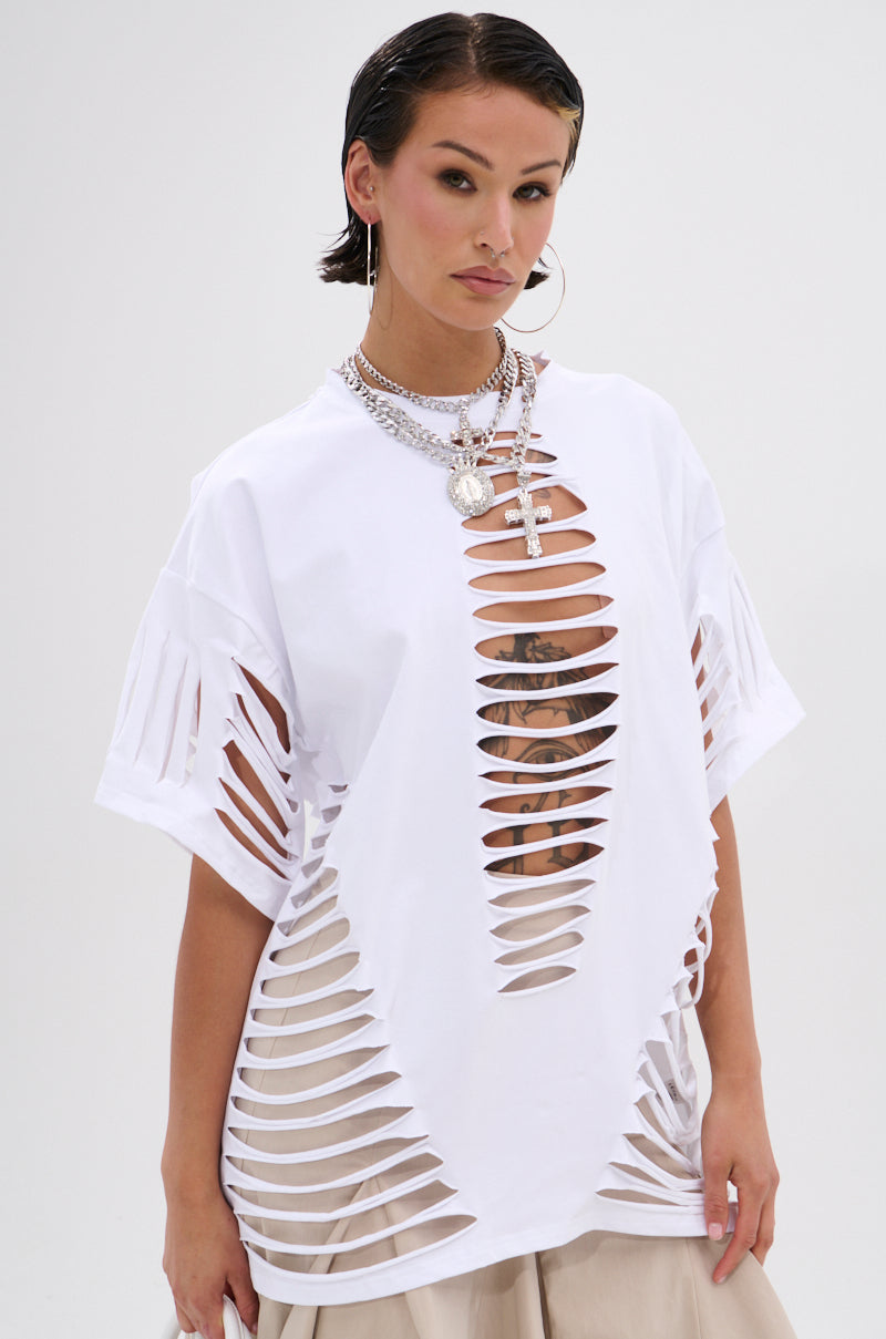 A woman in the SLASHED UP OVERSIZED SHORT SLEEVE TEE shows off a chest tattoo, styled with hoop earrings, layered silver necklaces, slicked-back short hair, and beige pants for an edgy look. 2