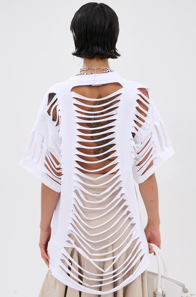 A person with short dark hair wears the SLASHED UP OVERSIZED SHORT SLEEVE TEE featuring horizontal slits down the back, revealing a tattoo. They pair it with a beige skirt and carry a white handbag. 4