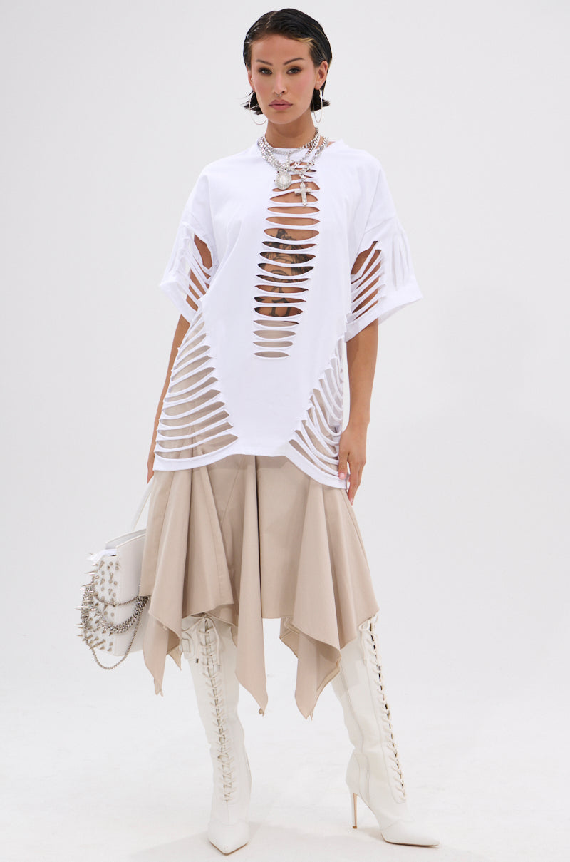 A woman poses against a white background wearing the SLASHED UP OVERSIZED SHORT SLEEVE TEE in white over a beige handkerchief-hem skirt, with knee-high lace-up boots, bold jewelry, and a studded handbag. 6