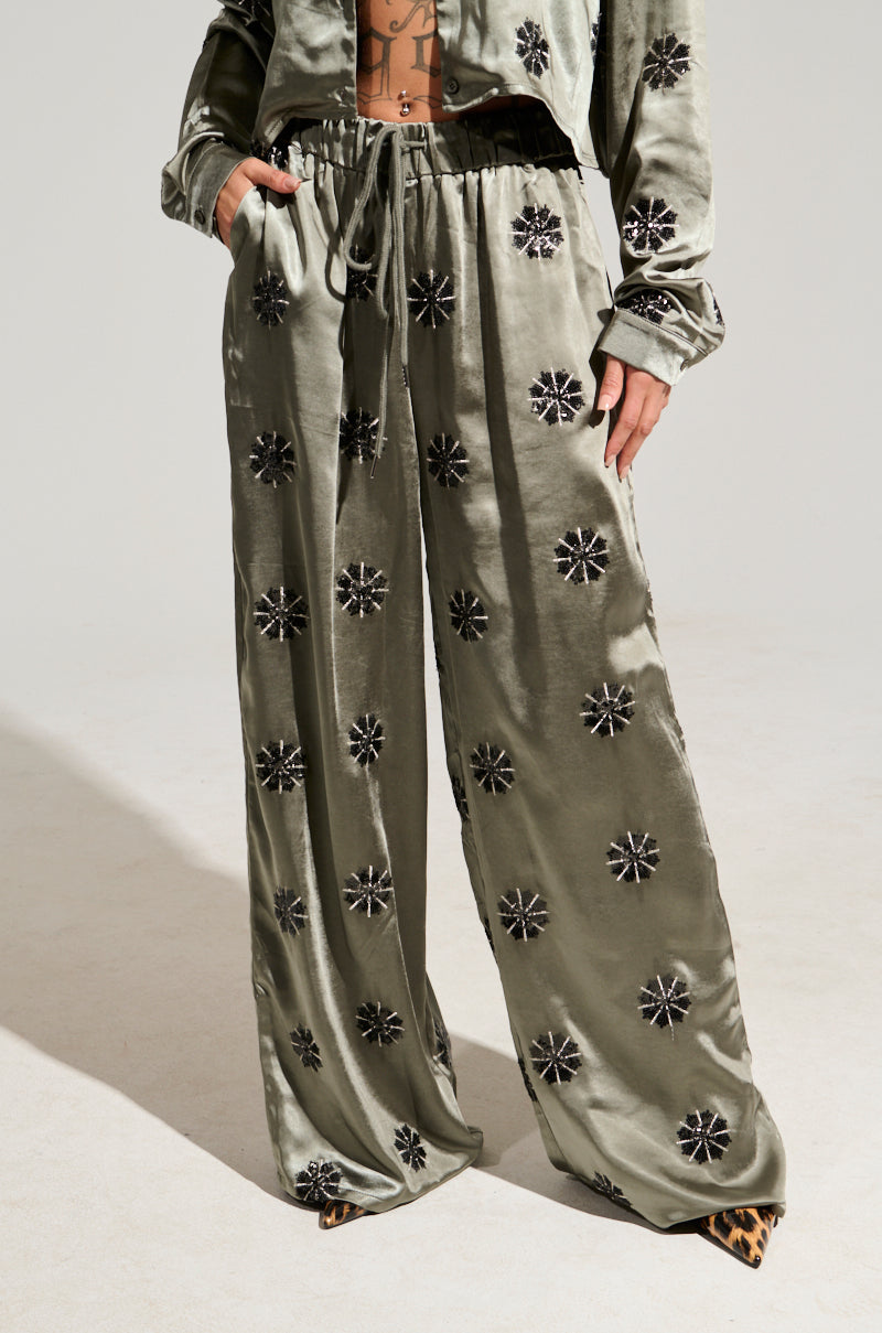 A person is dressed in a shiny, sage-green two-piece ensemble featuring long-sleeved tops and wide, flowy pants adorned with black floral patterns. The SUNSET DREAMS SATIN PANT IN SAGE showcases a high-rise elasticized waistband. They have their hands tucked into the pockets of the pants, while their shoes feature a striking leopard print design.