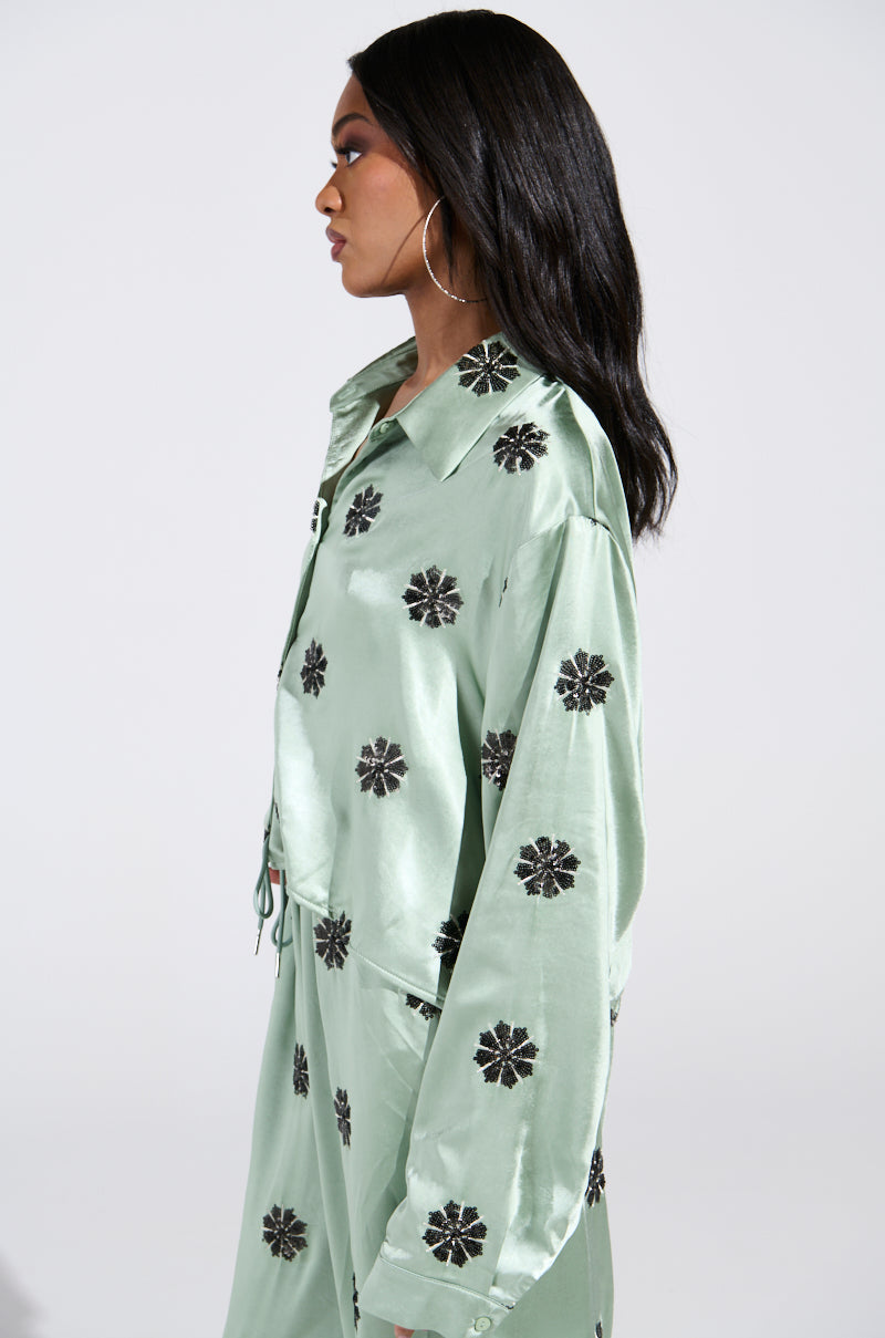 A person with long hair is in profile, facing left against a plain background, wearing the "DREAMS COMING TRUE SATIN BUTTON DOWN TOP" in light green with black flower patterns and sequin embellishments. They showcase large hoop earrings. 2