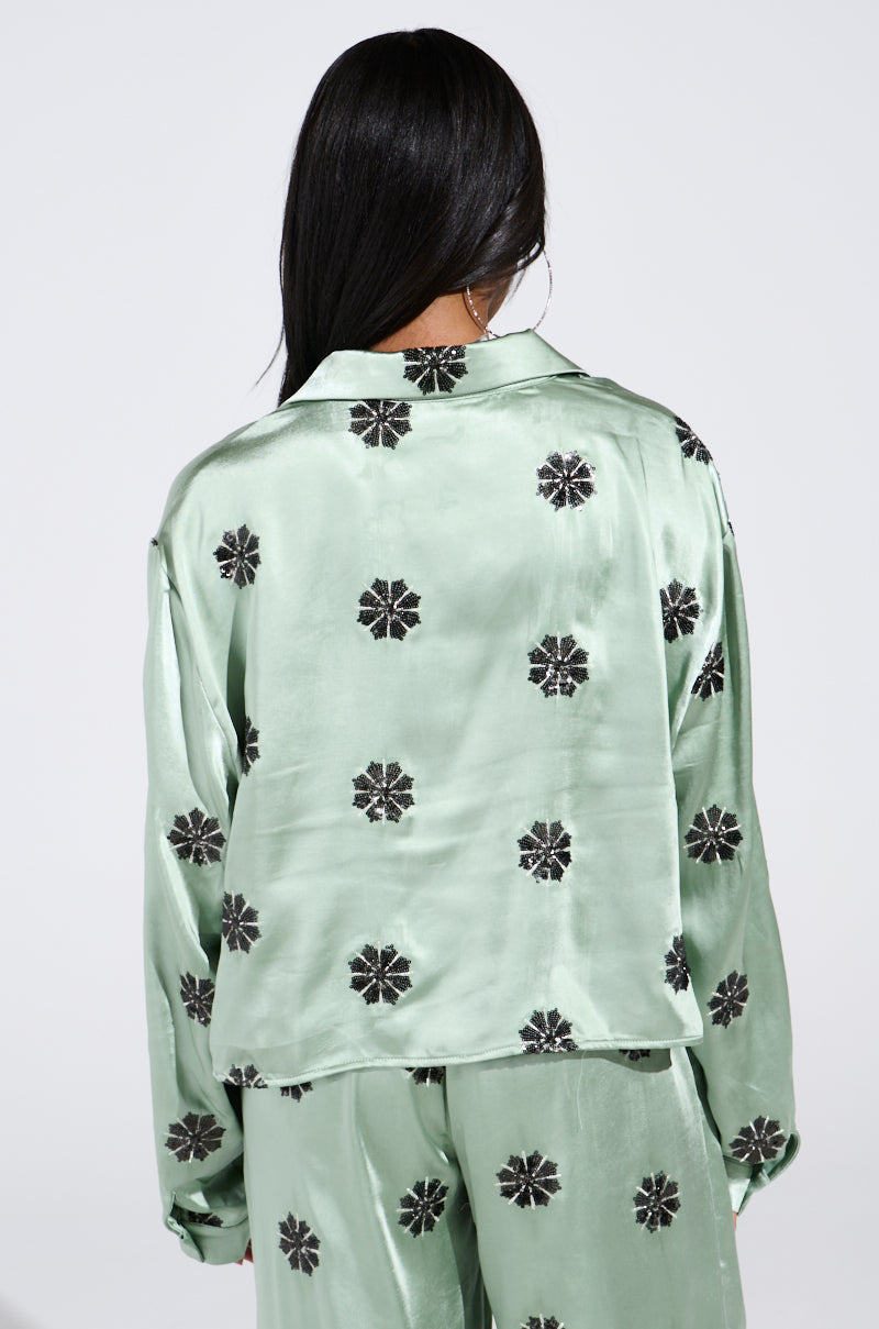 A person with long black hair is wearing a light green satin pajama set adorned with black geometric shapes and sequins. The AKIRA Label DREAMS COMING TRUE SATIN BUTTON DOWN TOP adds elegance as they turn away against a plain background. 3