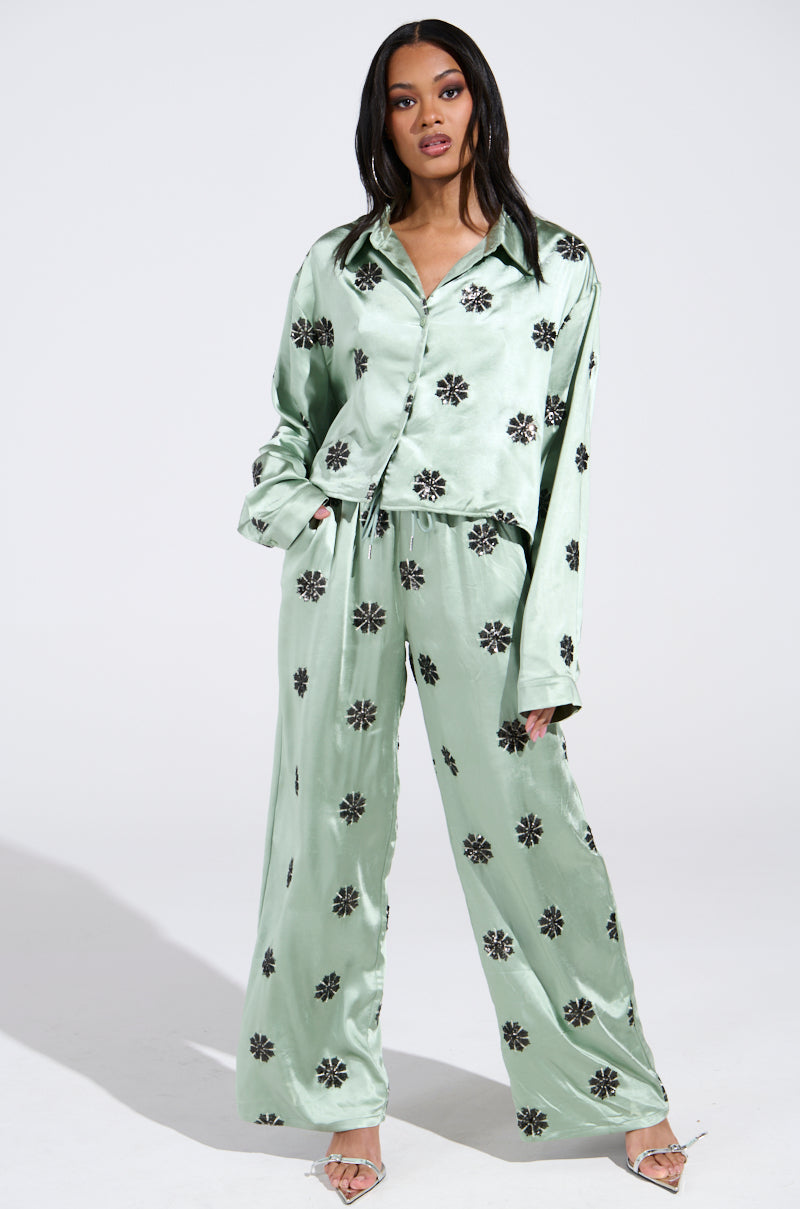 A woman models the DREAMS COMING TRUE SATIN BUTTON DOWN TOP and matching wide-leg pants in shiny, light green with dark floral patterns. She accessorizes with open-toed heels as her long, dark hair cascades down her shoulders against a plain backdrop. 4