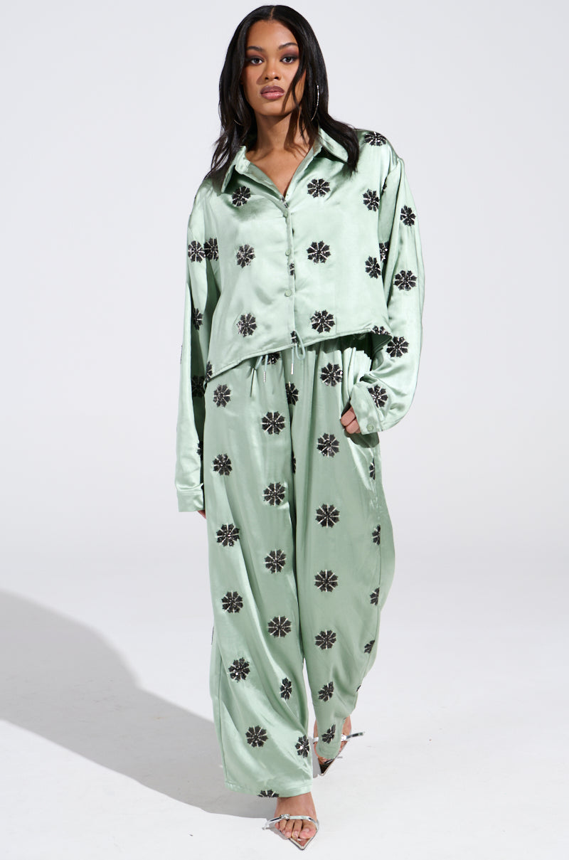 Dressed in an AKIRA Label Dreams Coming True ensemble, a person showcases a mint green satin pajama set with black floral patterns. The DREAMS COMING TRUE SATIN BUTTON DOWN TOP pairs elegantly with wide-leg pants against a neutral backdrop. 5