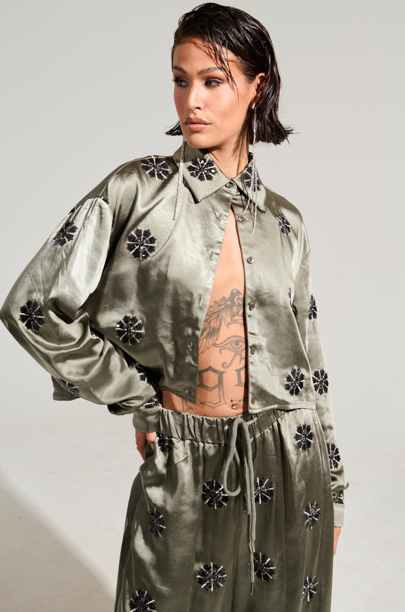 A person with short, wet hair models a shiny SUNSET DREAMS SATIN BUTTON DOWN TOP IN SAGE, featuring black floral patterns. The slightly open design reveals a tattoo on their torso as they pose with one hand on their hip against a plain background.
