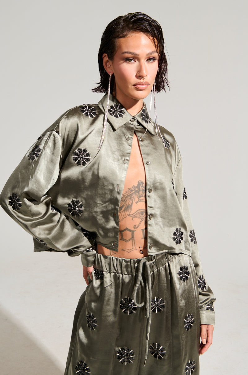 A person with short dark hair wears the "Sunset Dreams Satin Button Down Top in Sage" along with a matching skirt featuring black floral patterns. The shirt is left open to showcase a tattoo, and they complement their look with long, dangling earrings. The setting features a neutral gray background.