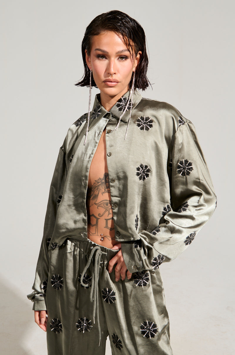 A person with short dark hair poses confidently in the open Sunset Dreams Satin Button Down Top in Sage, featuring black floral patterns that reveal a tattoo on their chest. The cropped boxy silhouette is accented by long, dangling earrings, and they maintain a neutral expression.