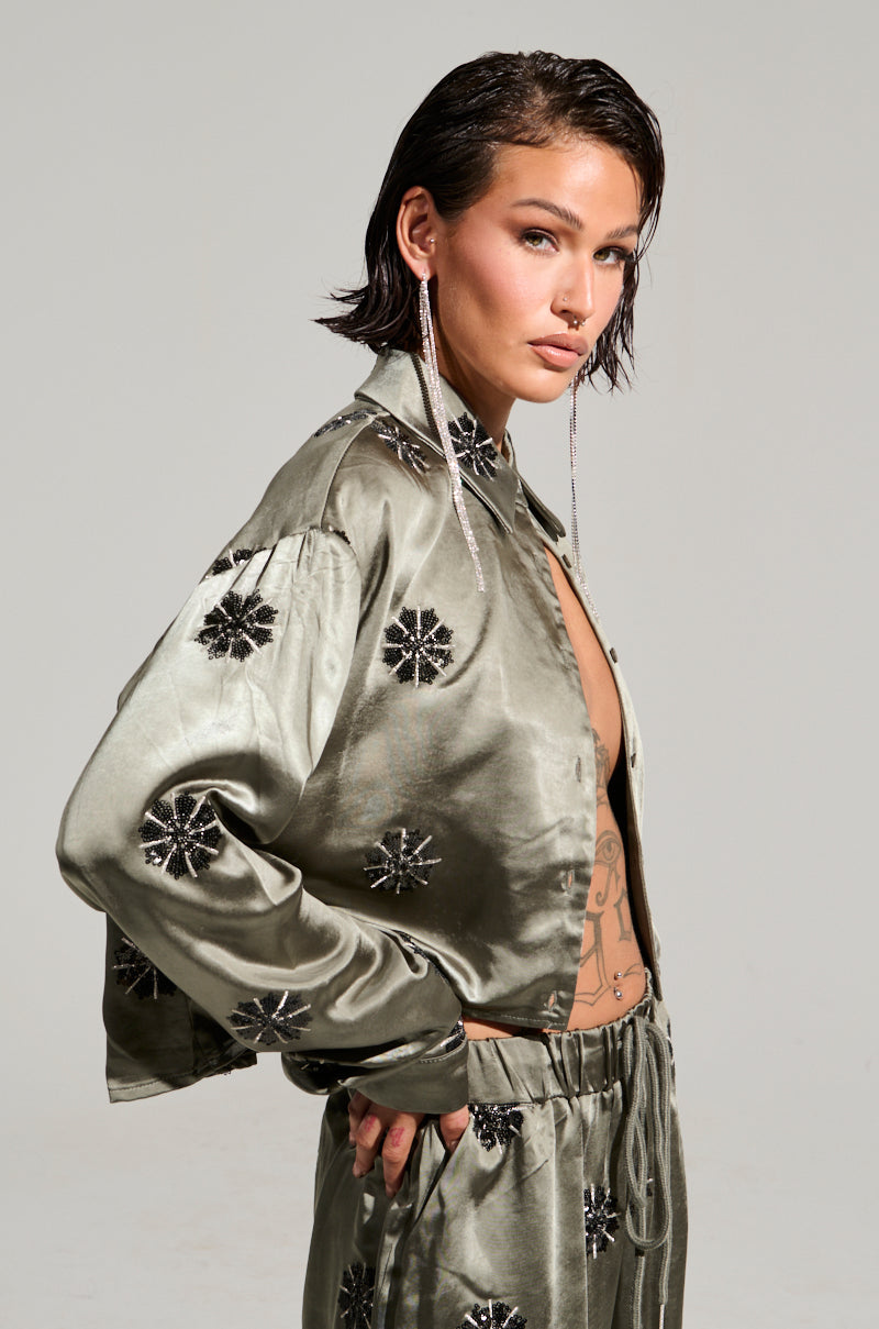 A person with short dark hair poses confidently in the SUNSET DREAMS Satin Button Down Top in Sage and a matching light olive green outfit adorned with black floral designs. The ensemble features a cropped boxy silhouette, enhanced by long, dangling earrings and a visible tattoo on their chest, set against a plain light gray background.