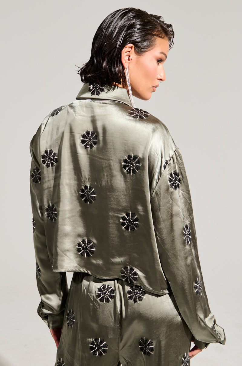 A person with short hair is wearing the Sunset Dreams Satin Button Down Top in sage, featuring a shiny olive green finish with black floral patterns. The ensemble includes a cropped boxy silhouette jacket and matching pants. The person gazes to the side, showcasing a long, dangling earring.
