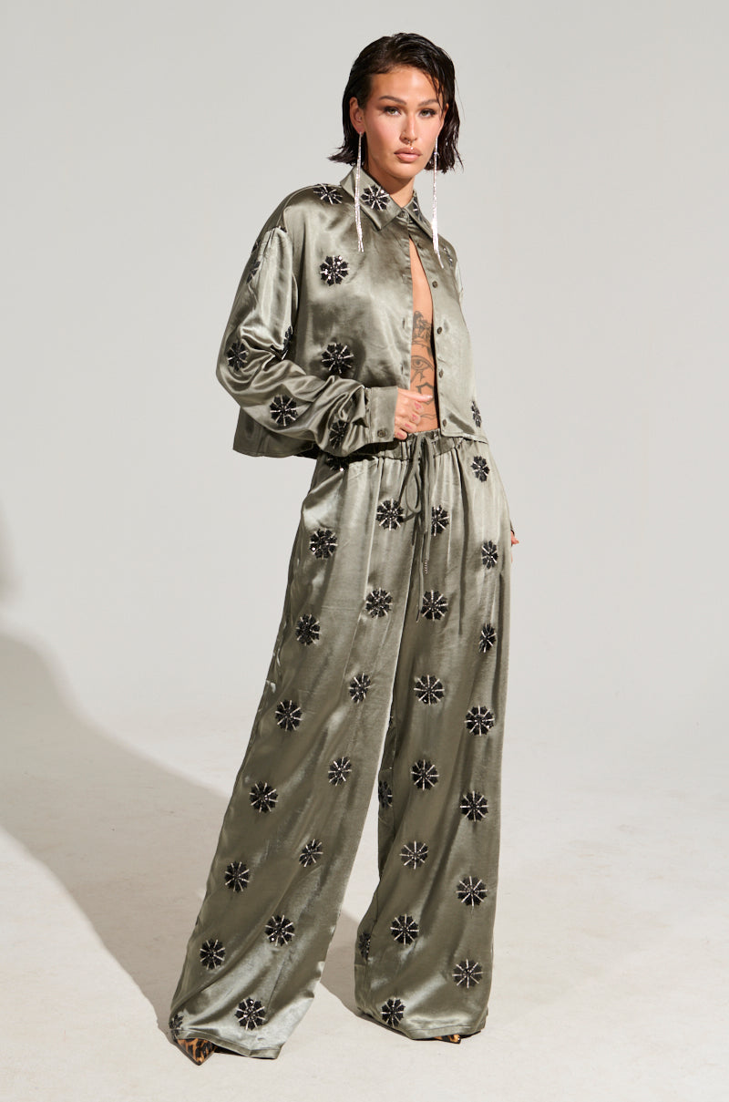 A person stands confidently wearing the SUNSET DREAMS Satin Button Down Top in a silky sage color, part of a stylish two-piece ensemble featuring wide-leg pants with floral embroidery. They have short brown hair and are posing against a plain background.