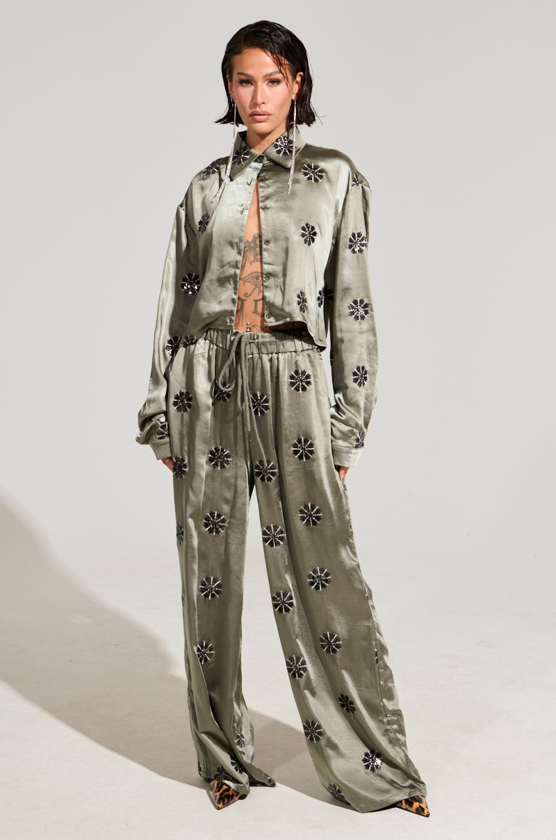 A person poses confidently in the SUNSET DREAMS Satin Button Down Top in sage, featuring a shiny, olive-green hue with black floral patterns. The outfit boasts a cropped boxy silhouette paired with wide-leg pants, all accentuated by high heels.