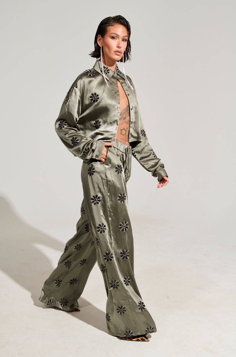 A person with short hair walks confidently, wearing the Sunset Dreams Satin Button Down Top in a sage hue. Paired with wide-leg pants, this chic ensemble features sequins and a floral pattern, presenting a stylish boxy silhouette. The backdrop is kept plain and light-colored.