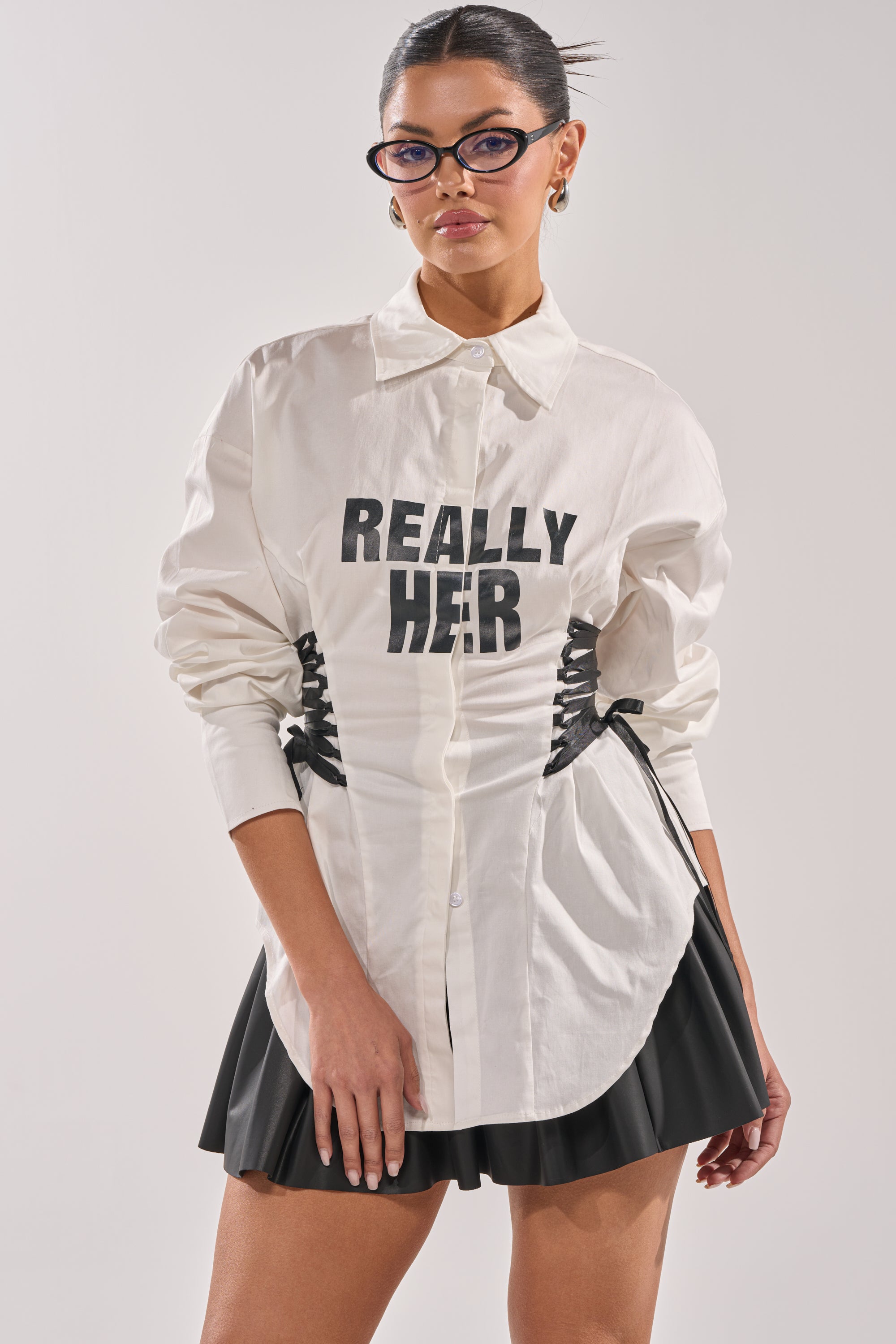 A woman wearing glasses, hoop earrings, and the REALLY HER POPLIN BLOUSE styled as a white button-down with "REALLY HER" on the front, paired with a black pleated mini skirt, poses confidently against a plain background. 0