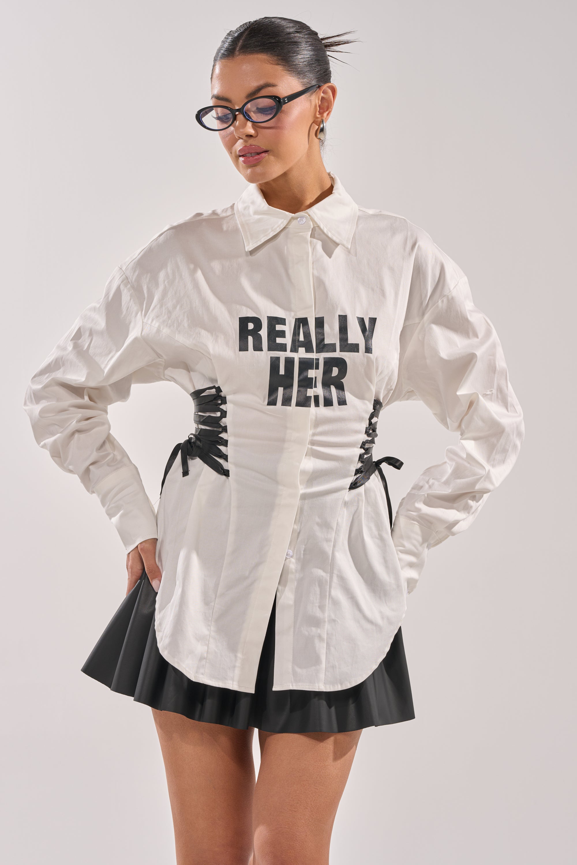 A woman in glasses wears the REALLY HER Poplin Blouse—a white button-up with “REALLY HER” text and black lace-up sides—paired with a pleated black mini skirt, standing hands on hips against a plain background. 1
