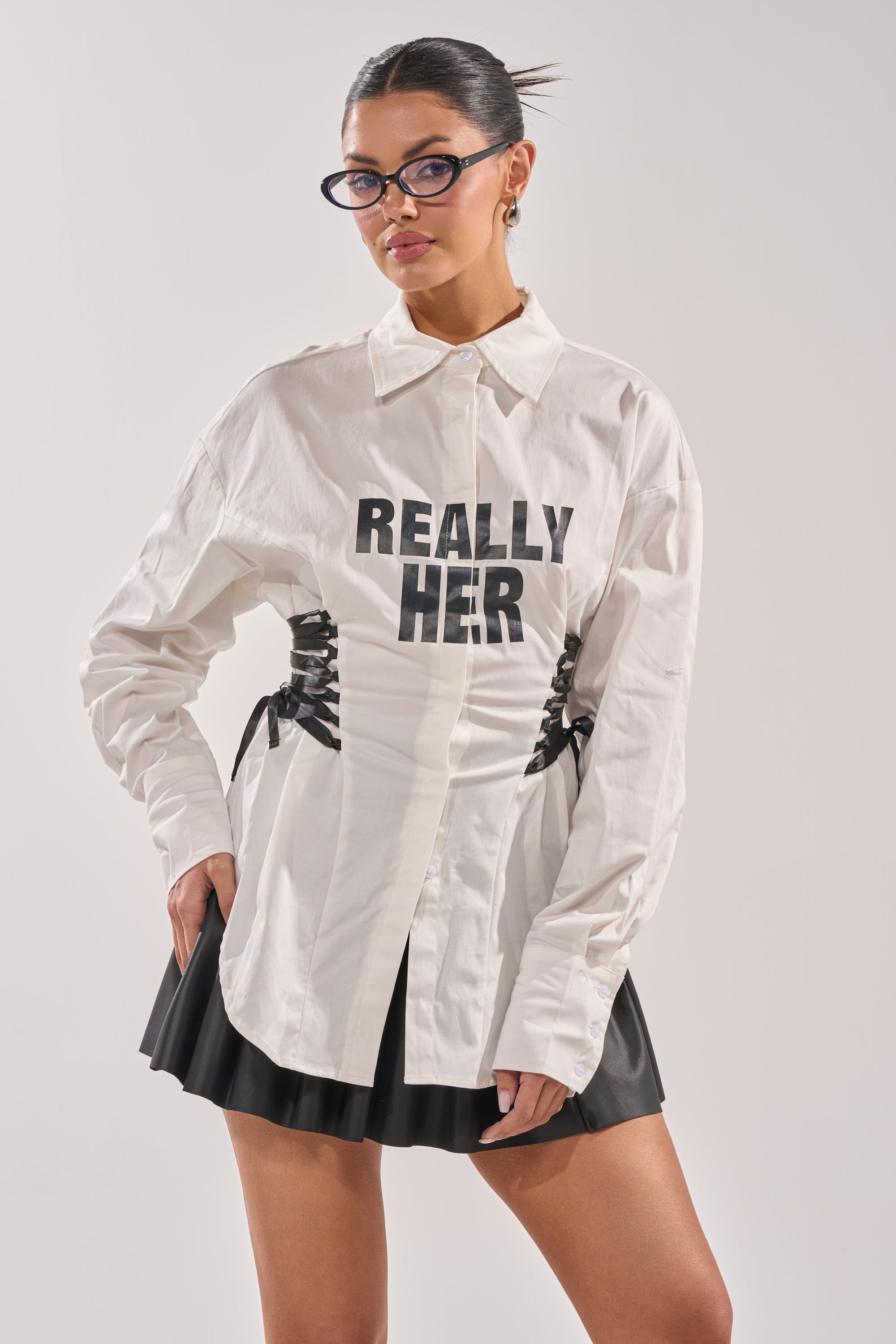A person with dark hair in a sleek bun wears glasses, the REALLY HER POPLIN BLOUSE, and a black pleated skirt, posing confidently with one hand in their pocket. 2