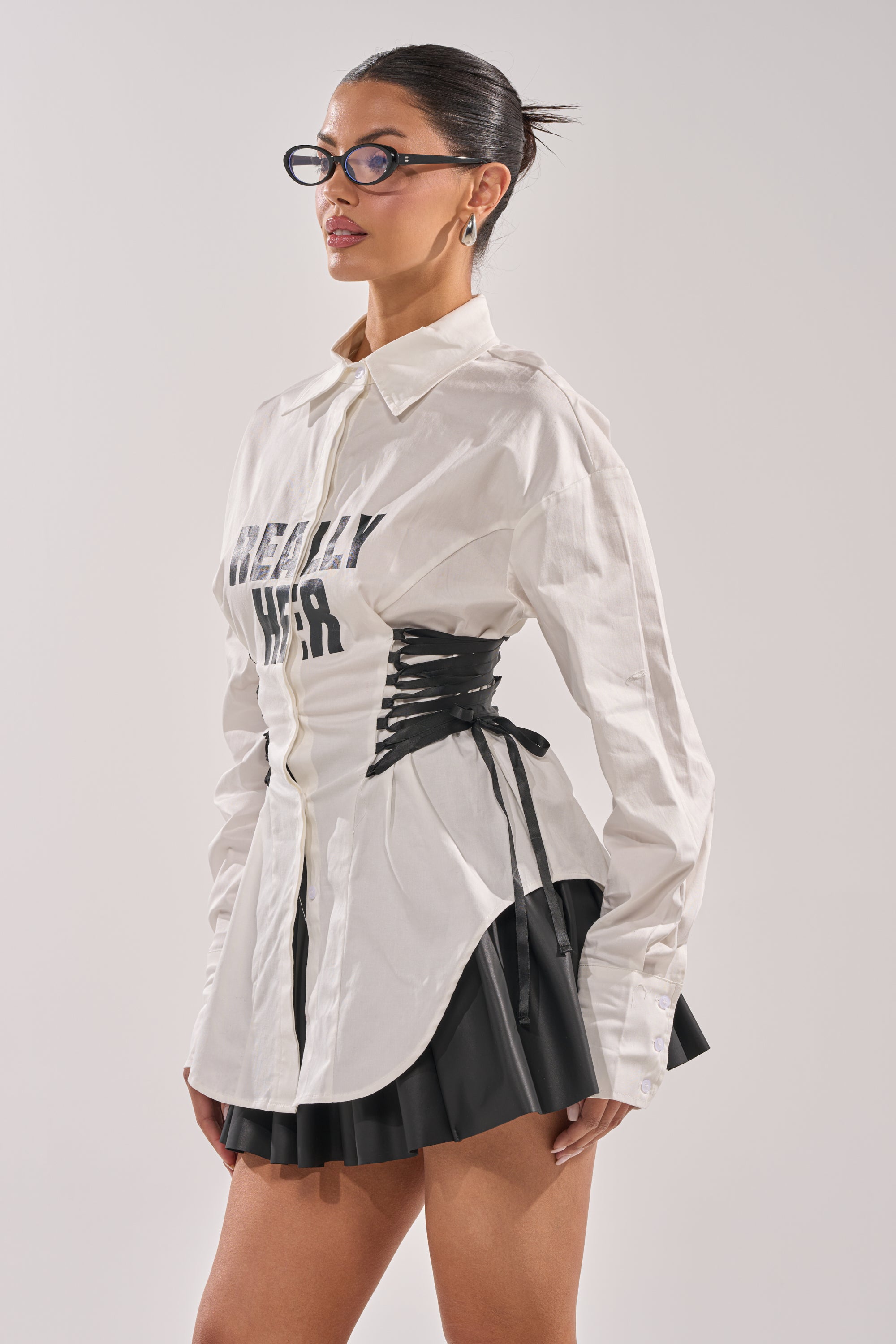 A woman with slicked-back hair and glasses styles the REALLY HER POPLIN BLOUSE, cinched at the waist with black lacing, paired with a black pleated mini skirt for a chic look against a plain background. 3
