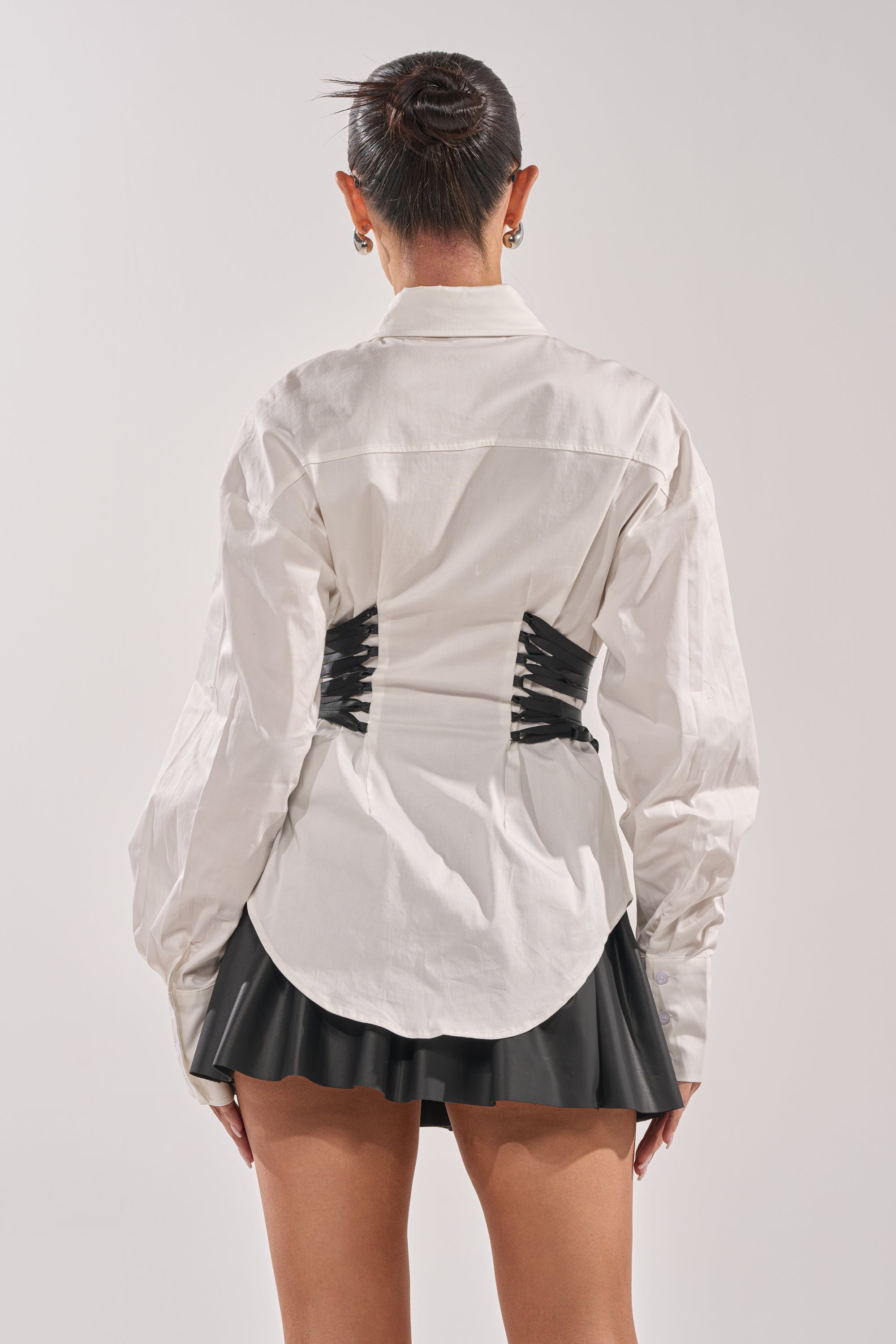 A person faces away, wearing a white REALLY HER POPLIN BLOUSE with black corset-style detail at the waist and a short black pleated skirt. Their hair is in a neat bun. The background is plain and light-colored. 4