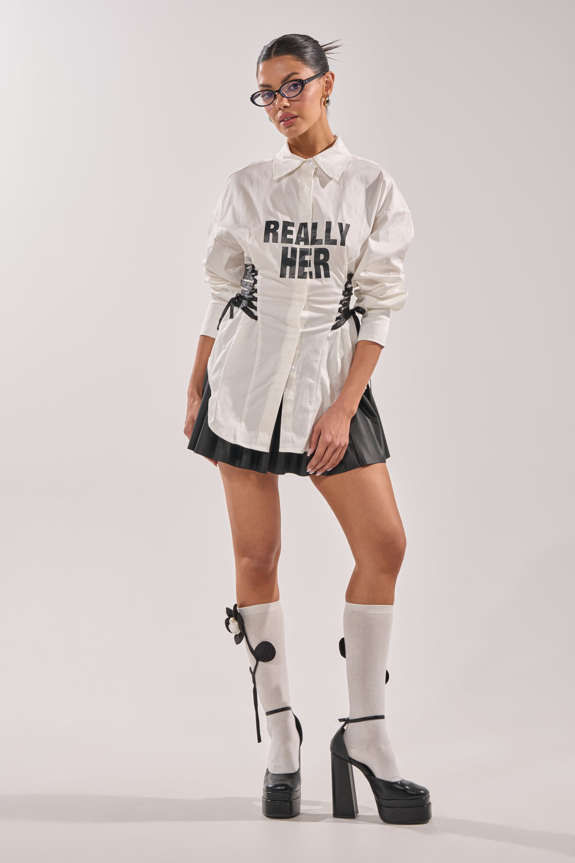 A woman stands against a plain background wearing black glasses, the REALLY HER POPLIN BLOUSE, a black pleated miniskirt, white knee-high socks with black dots, and black platform heels. 6
