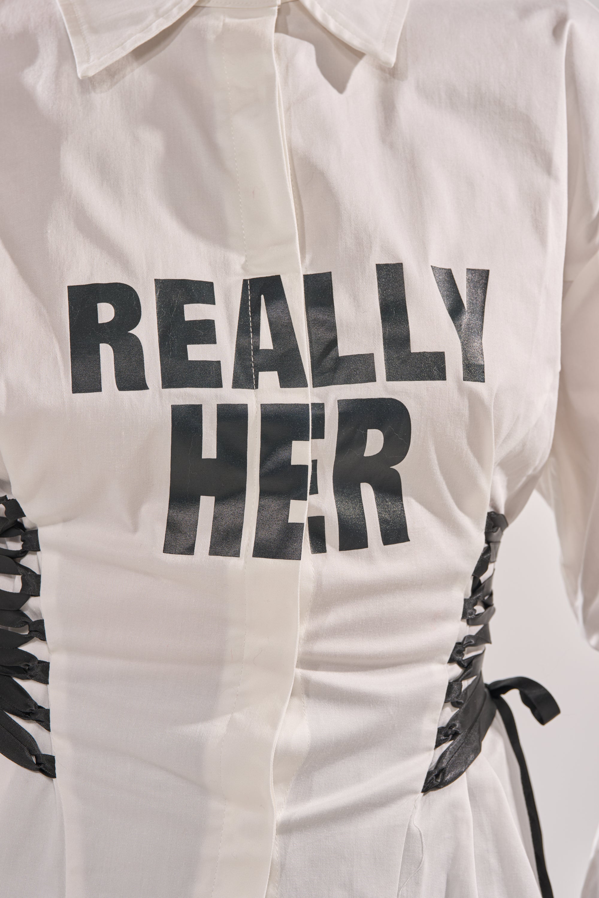 A close-up of the REALLY HER POPLIN BLOUSE, a white lace-up corset style top with bold "REALLY HER" lettering. Black side laces cinch the waist, making it a standout choice for a statement night-out look. 8