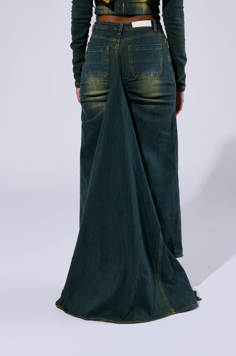 A person is shown from the back, clad in the Y2K-inspired BEFORE YOU GO DENIM SKIRT. The garment boasts a dramatic, high-rise cut with a long tail that elegantly drapes to the floor from the back of the waist, offering a unique and striking design element. The dark blue denim fabric features subtle faded patches for added character.