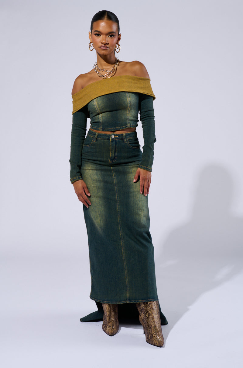 A woman stands confidently against a plain background, donning the BEFORE YOU GO OFF THE SHOULDER DENIM TOP with a mustard yellow finish. Her Y2K-inspired outfit includes a high-waisted long denim skirt and snakeskin boots. She accessorizes with large hoop earrings and layered necklaces, exuding effortless style.