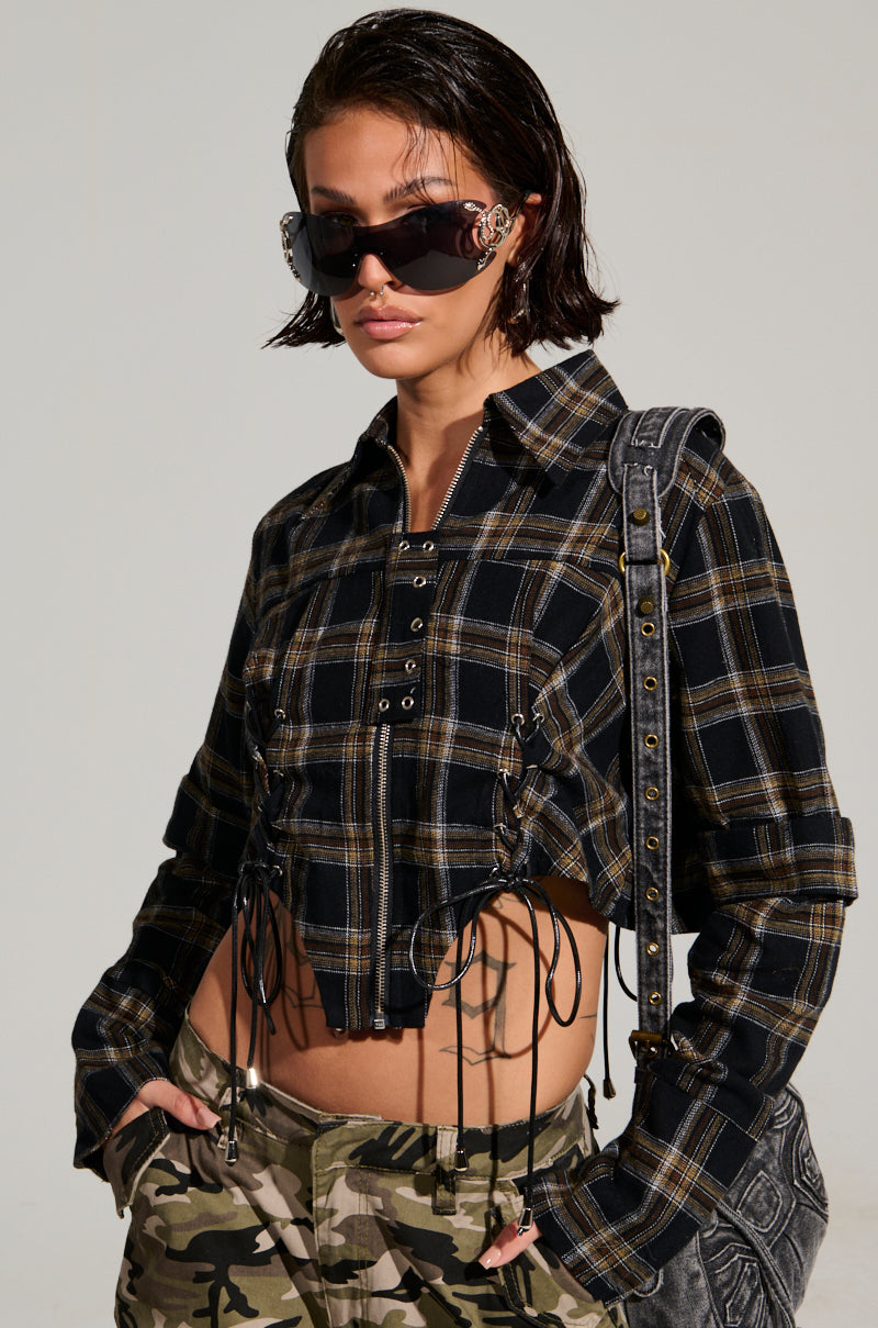 A person with short brown hair wears large sunglasses and the UNSPEAKABLE BUTTON DOWN PLAID SHIRT in black and yellow, paired with camouflage pants. They carry a black shoulder bag and stand casually with their hands in their pockets against a neutral background.