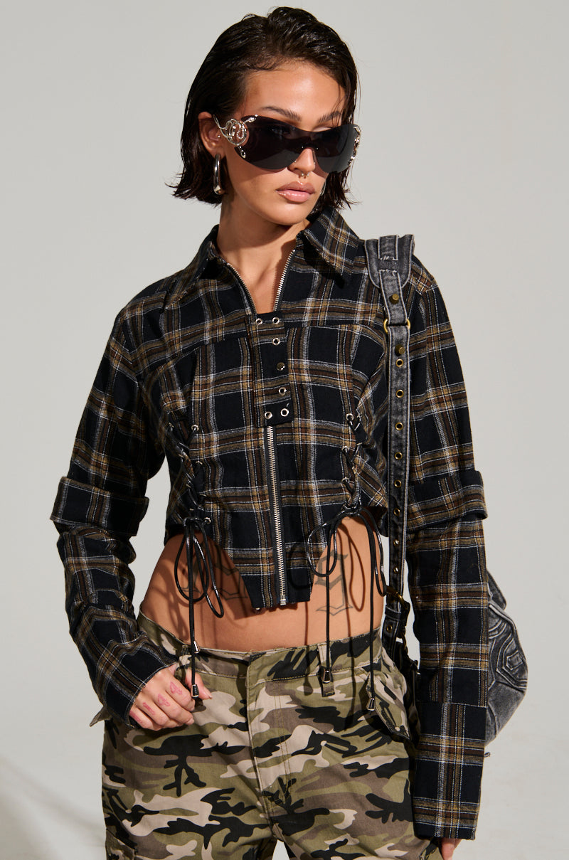A person with short black hair wears oversized sunglasses and a cropped UNSPEAKABLE BUTTON DOWN PLAID SHIRT in black and yellow with lace-up details, accompanied by camo pants. They carry a gray backpack over one shoulder.