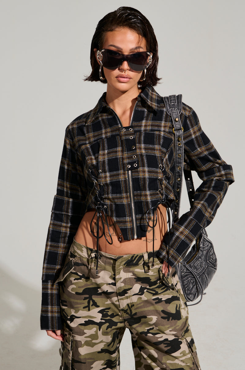 A person wearing the UNSPEAKABLE BUTTON DOWN PLAID SHIRT, featuring a cropped silhouette and lace-up details, paired with camouflage cargo pants. They accessorize with large black sunglasses and a textured shoulder bag, posing against a neutral background.
